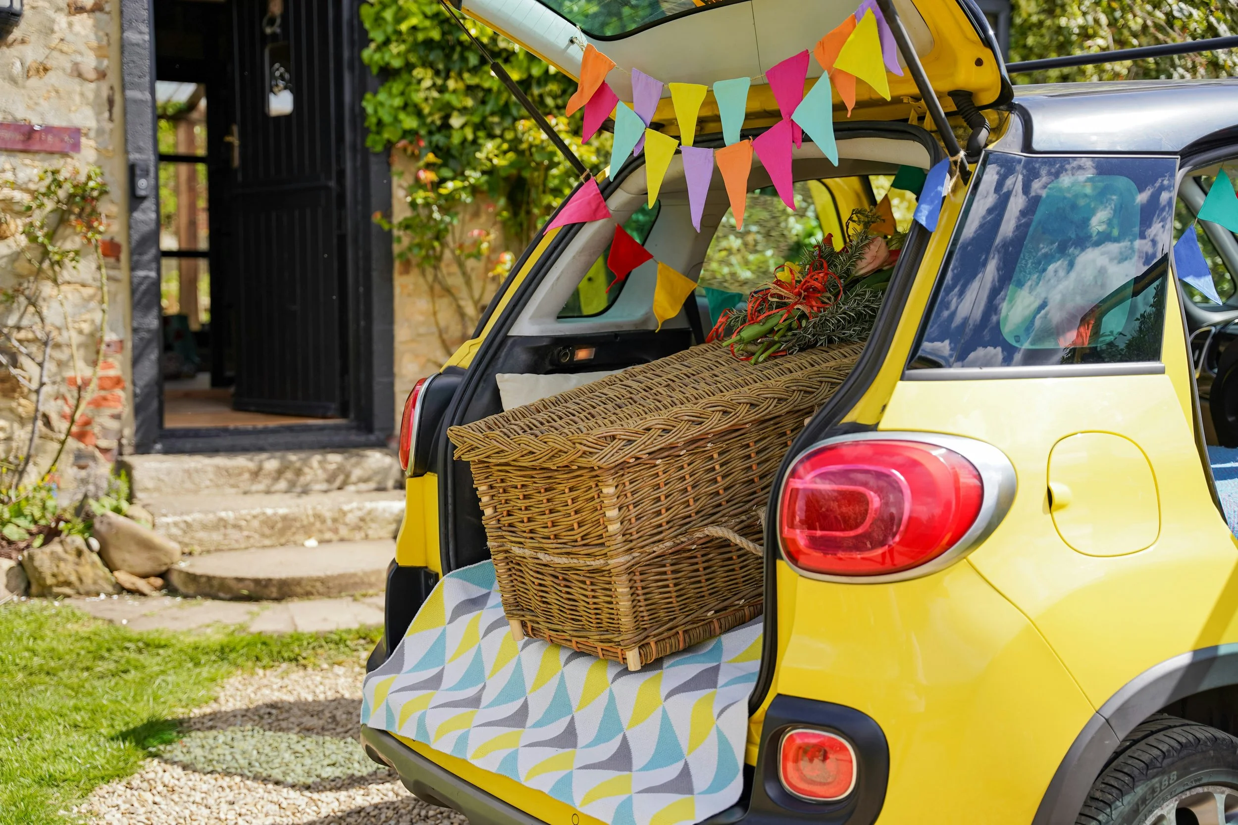 A yellow compact car decorated with colorful bunting, parked in front of a stone building with an open gate. The open trunk contains a wicker chest, a flower bouquet, and a pillow, with a patterned cloth underneath.