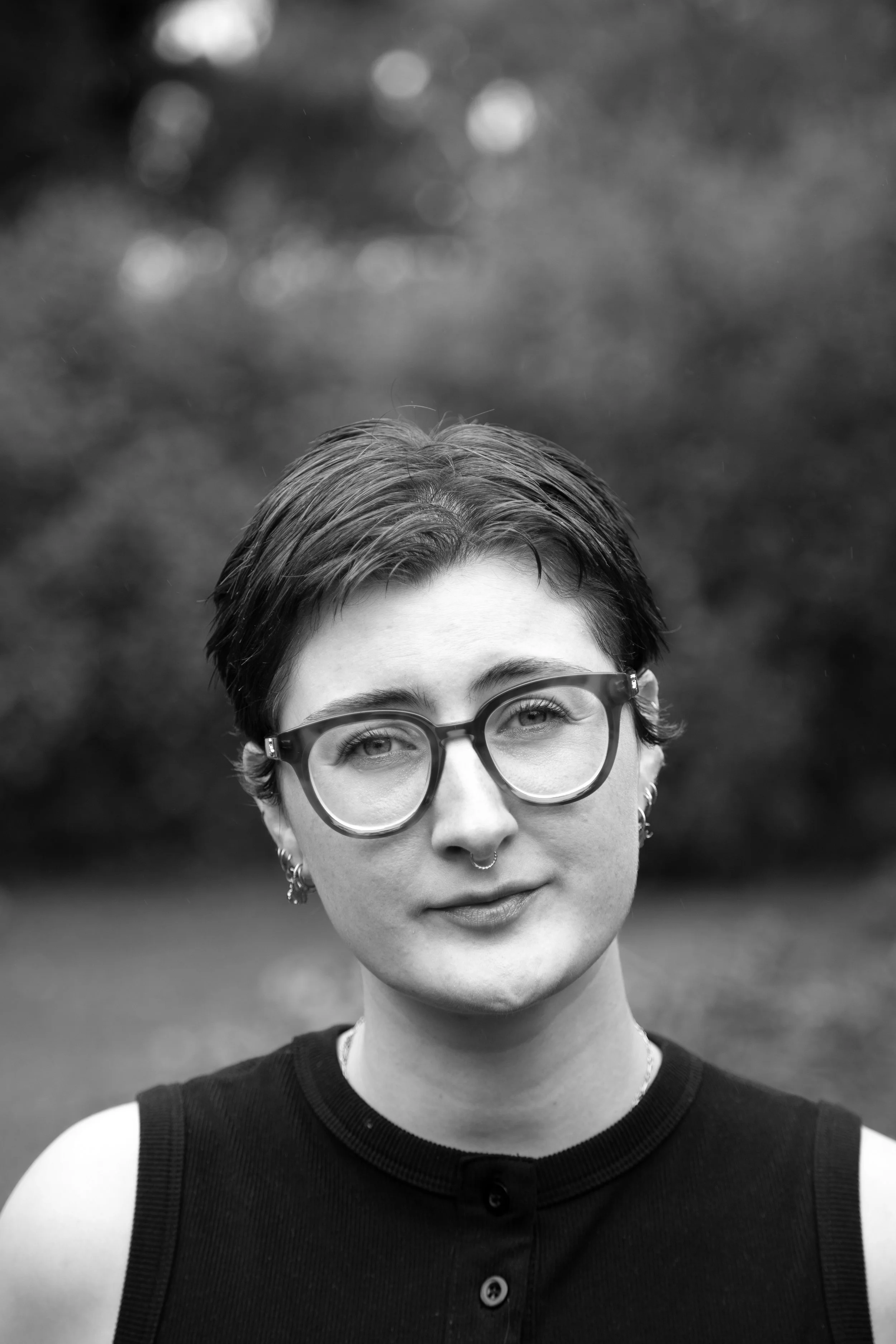 Black and white photo of a young person with short hair, glasses, earrings, and a septum piercing, wearing a sleeveless top, outdoors with blurred trees in the background.