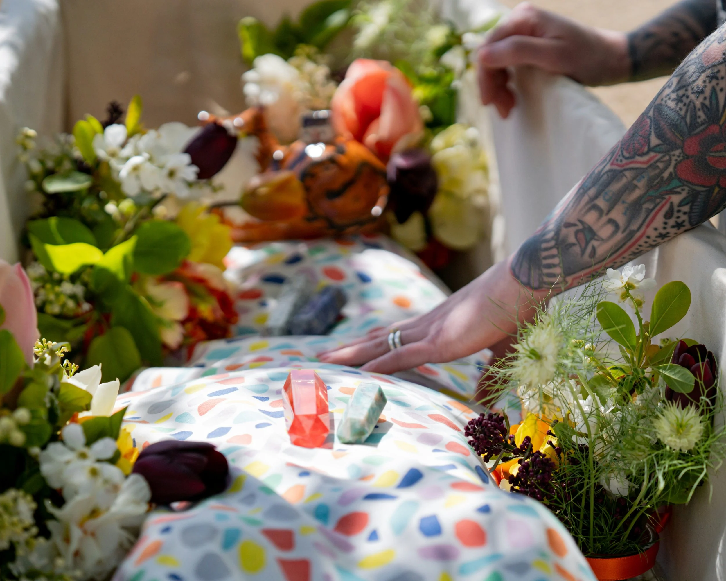 A person with tattoos on their arms placing their hands on a colorful blanket covered with various types of flowers and small crystal stones.