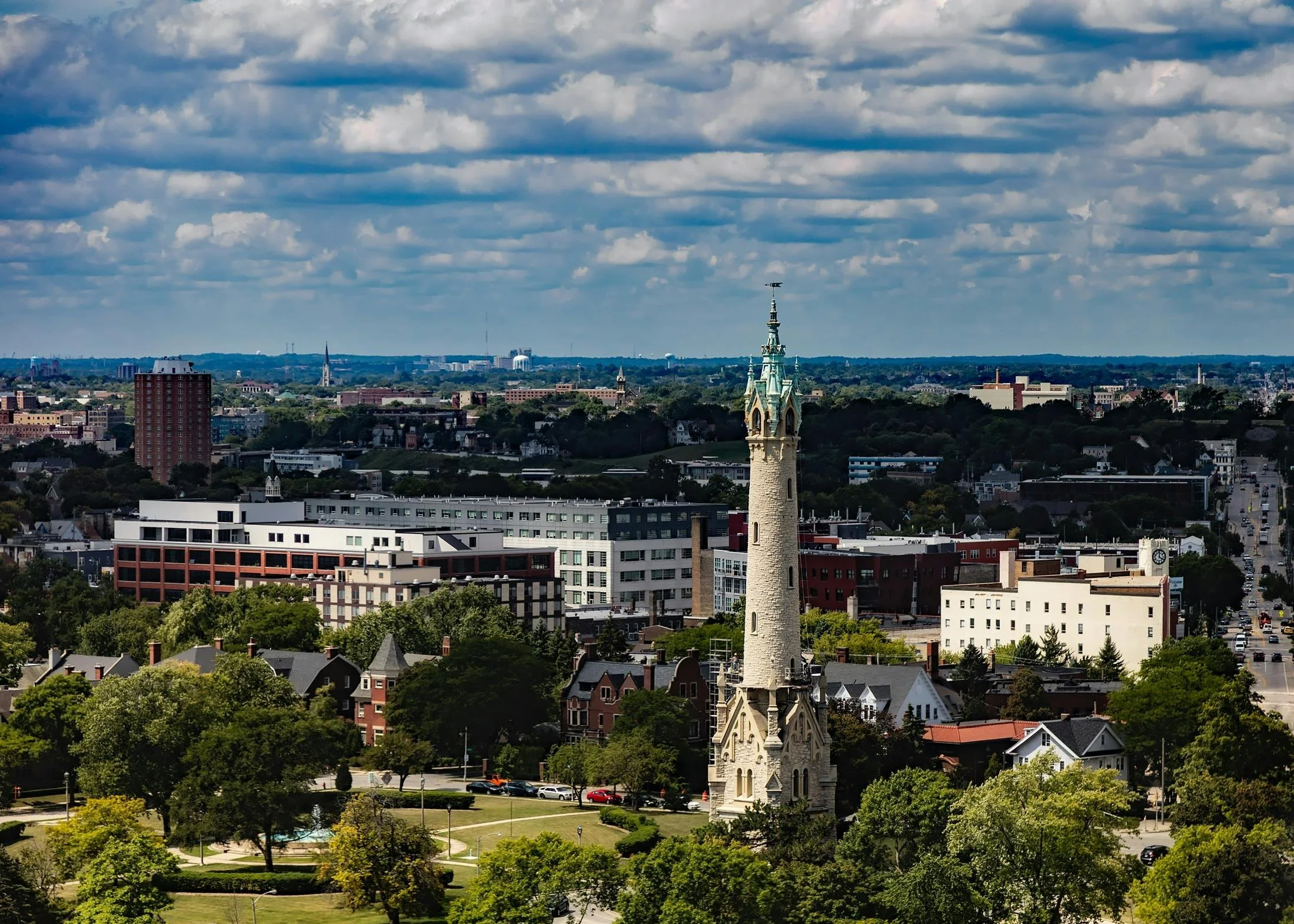 Elevated view of a city neighborhood featuring a tall stone clock tower rising above trees, homes, and mid-rise buildings under a partly cloudy blue sky.