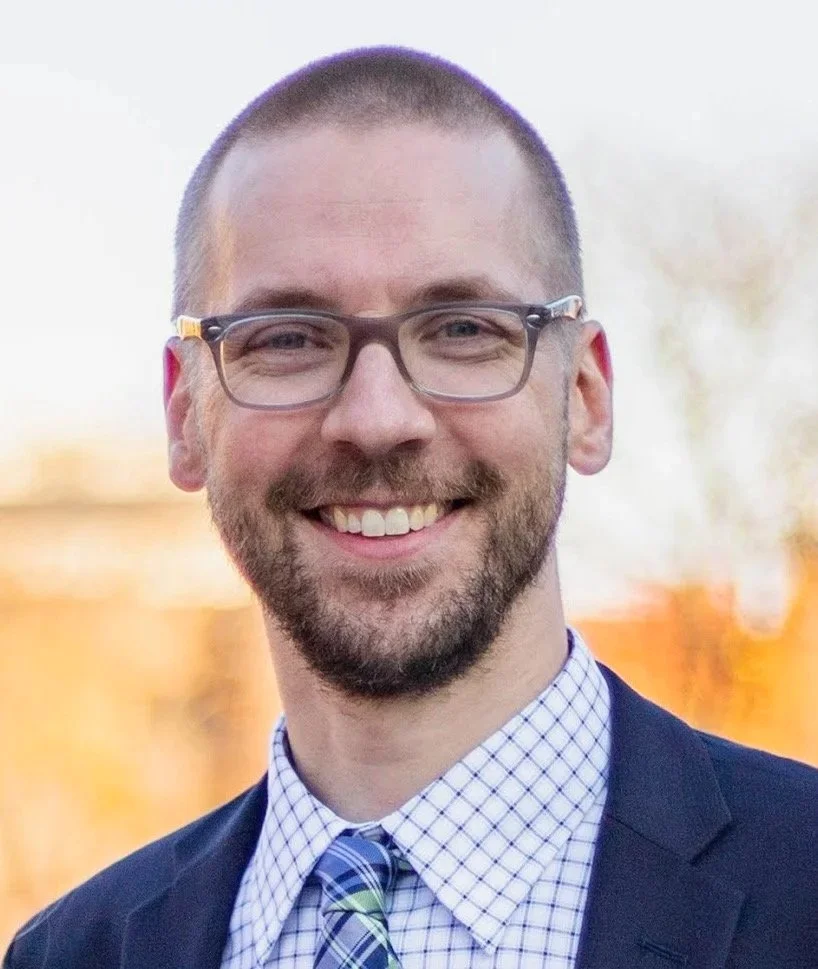 Portrait of Colin Lavey smiling outdoors, wearing glasses, a navy blazer, a checkered dress shirt, and a patterned tie, with a softly blurred background.