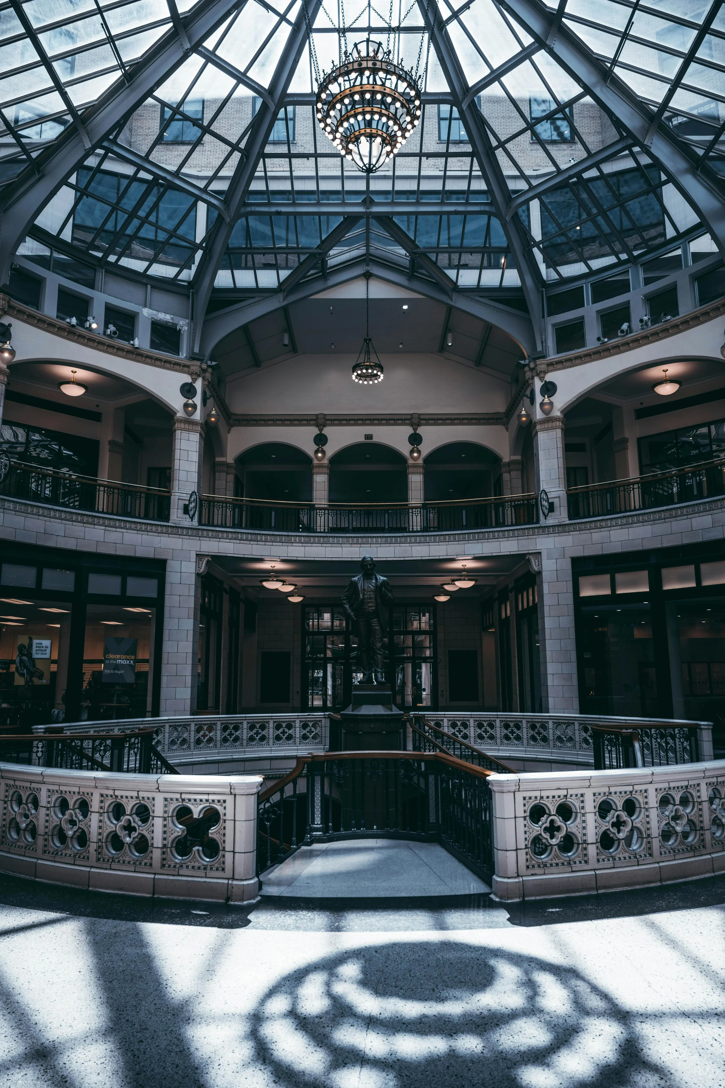 Sunlit interior of Plankinton building with a glass skylight ceiling, ornate railings, hanging chandeliers, and a bronze statue centered on the ground floor.
