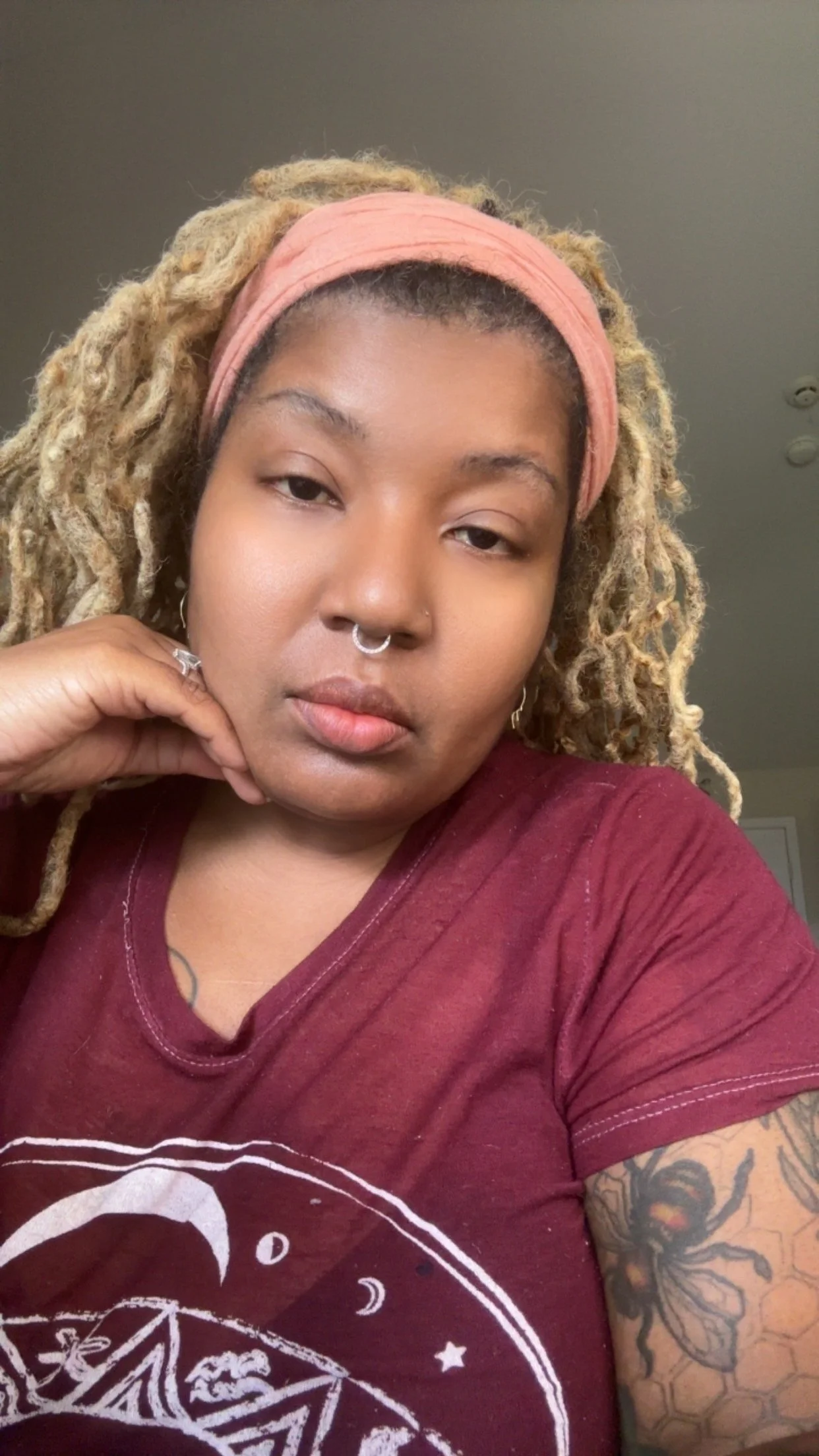 A young woman with blond dreadlocks wearing a pink headband and a maroon T-shirt, resting her chin on her hand, with visible tattoos on her arm.