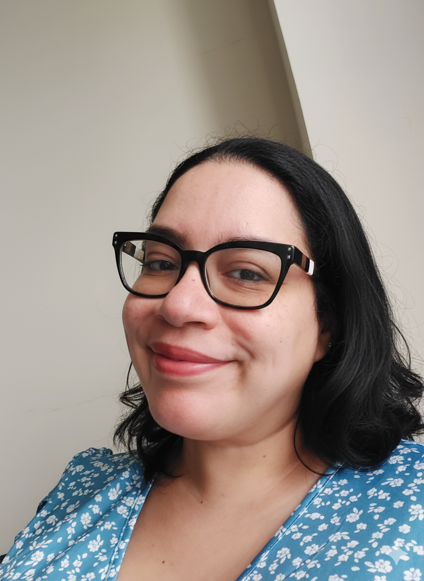 A woman with black hair and glasses smiling for a selfie, wearing a blue floral top, in front of a plain wall.