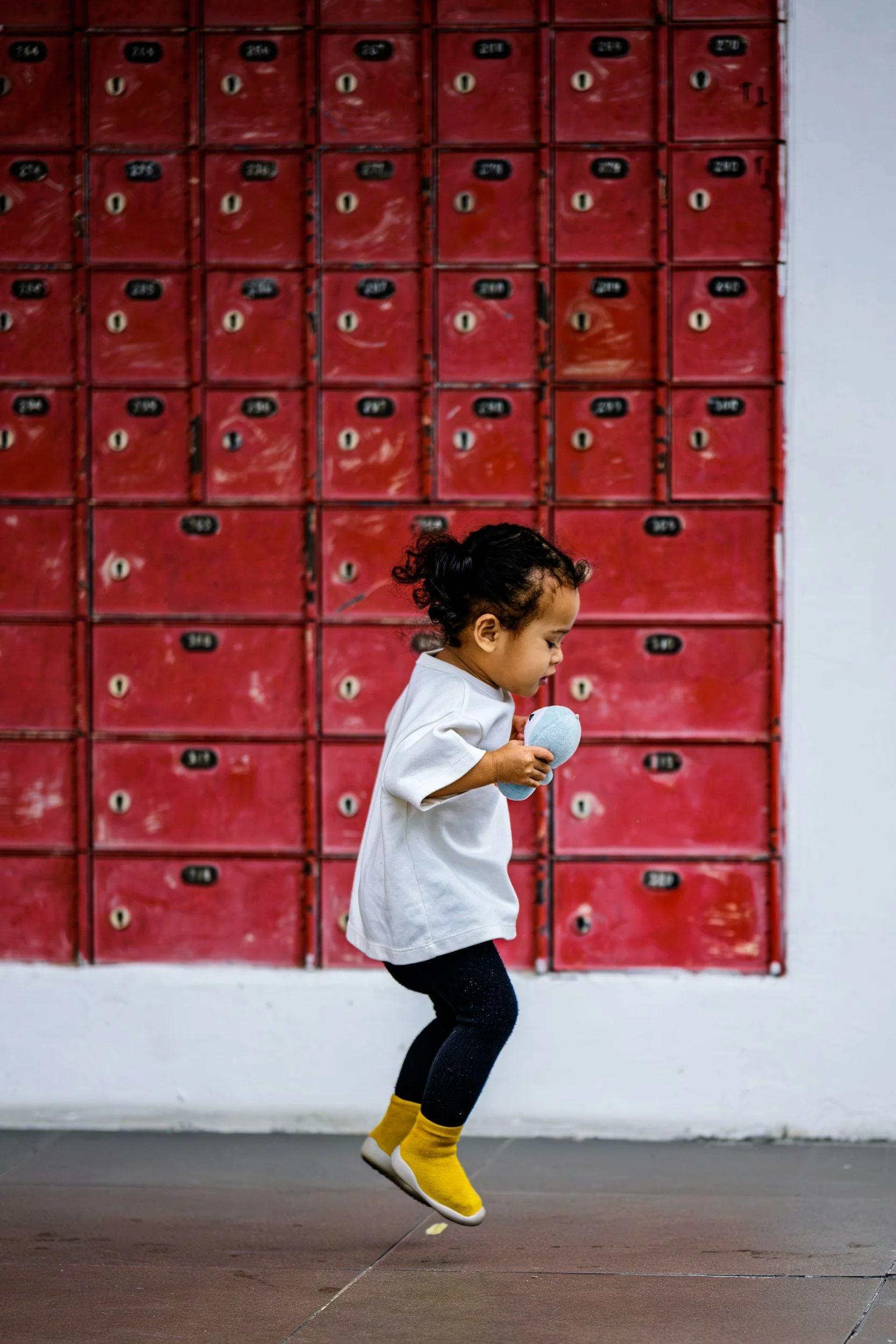 A young girl jumping in front of red lockers, holding a blue ball, dressed in a white shirt, dark pants, and yellow socks.