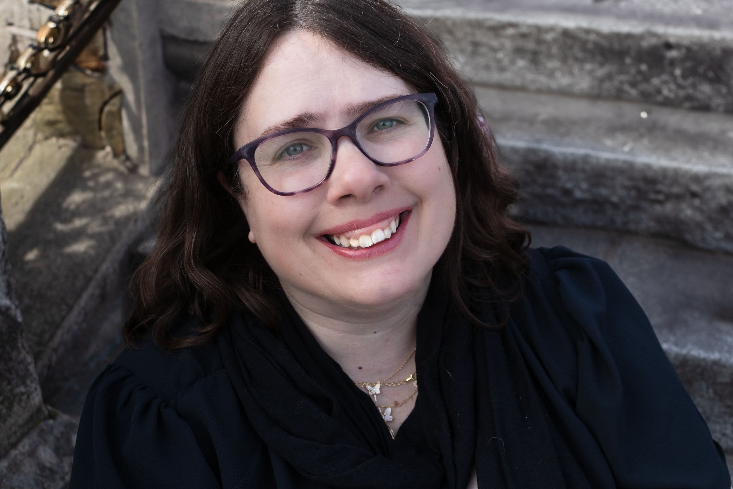 A picture of Tammie Meloy- woman with glasses and dark brown wavy hair smiling for a photo outdoors, sitting on stone steps.