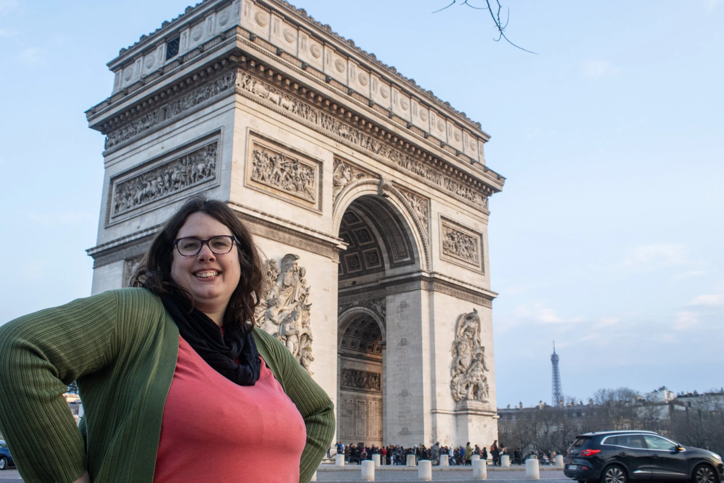 Tammie Meloy- A smiling woman with glasses stands in front of the Arc de Triomphe in Paris, France, with the Eiffel Tower visible in the background.