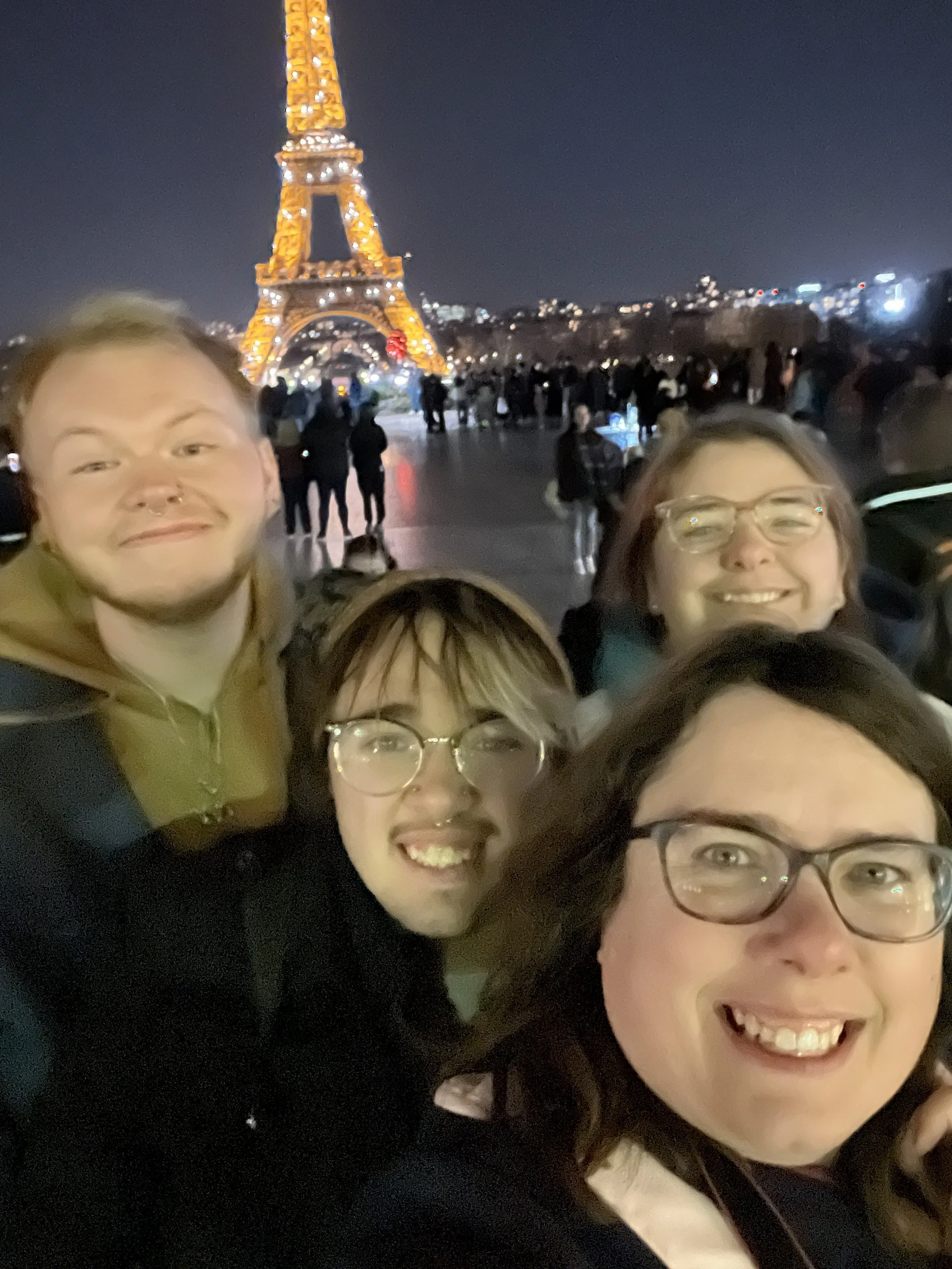 Tammie and family taking a selfie at night with the Eiffel Tower illuminated in the background in Paris.