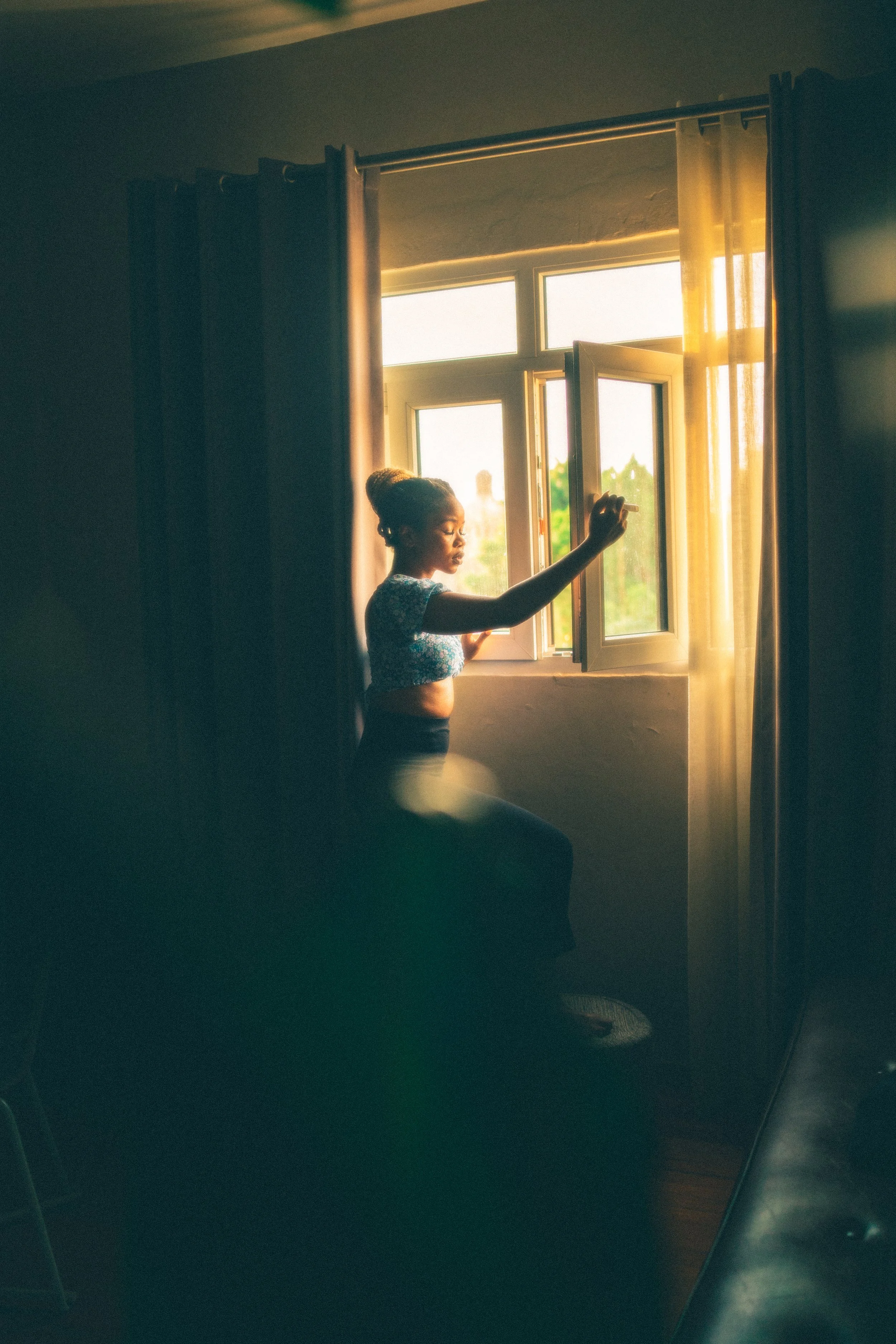 A woman sitting by an open window, bathed in warm sunlight, with curtains on either side of the window.