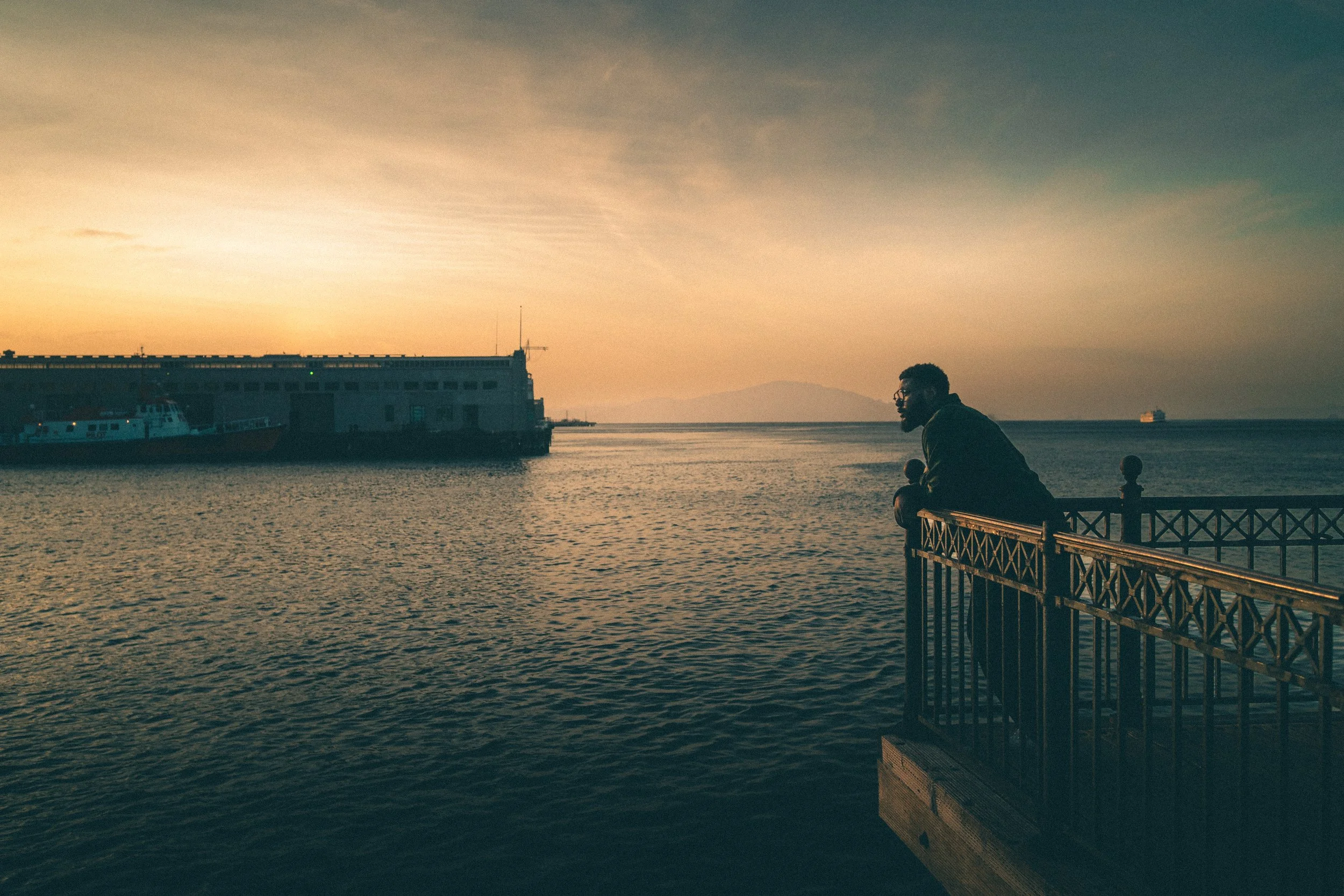 A man leaning on a railing by the water at sunset, with boats and a large building in the background.