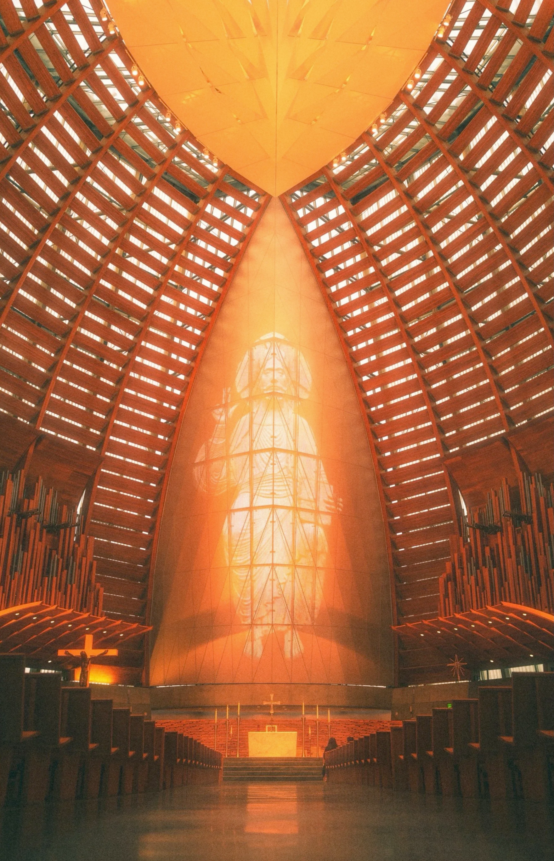 Interior of a church with a large illuminated crucifix at the front, wooden pews, and a tall, pointed, colorful stained-glass window depicting Jesus Christ.