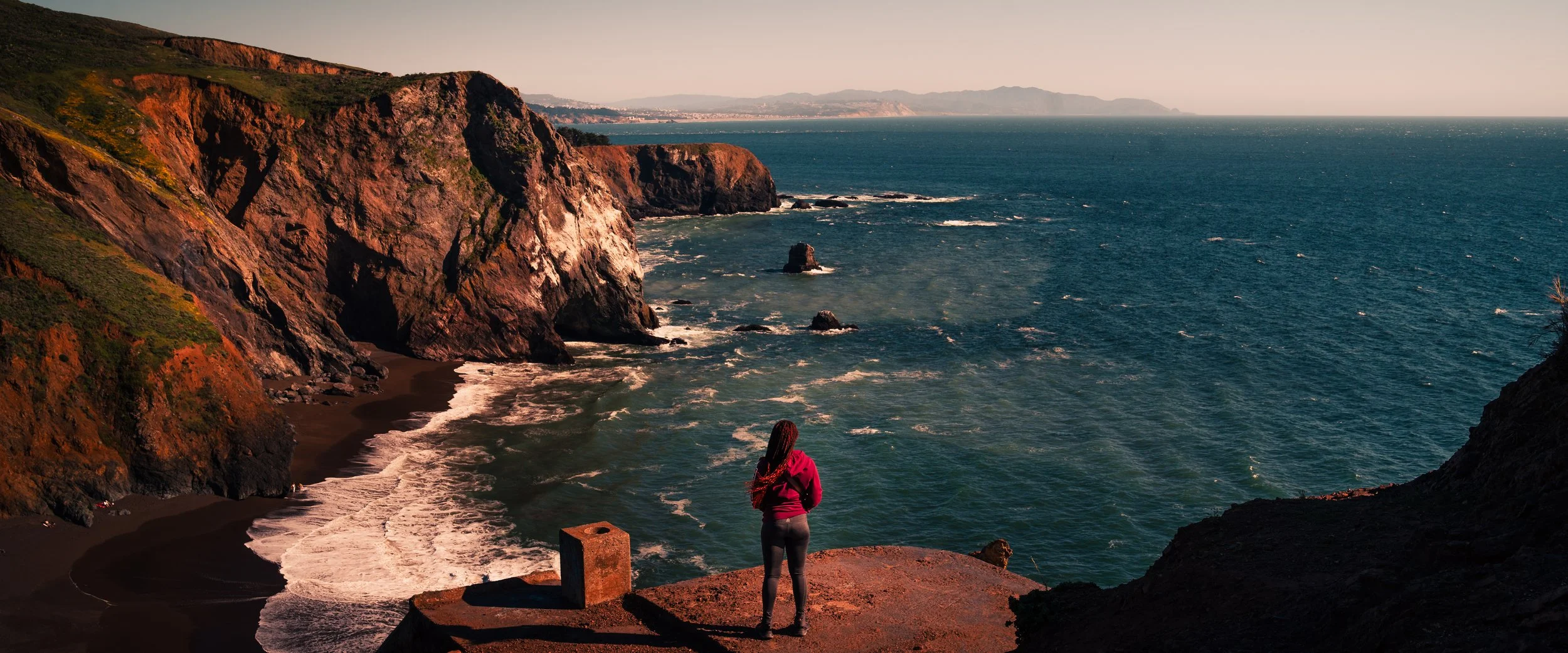 A woman in a pink jacket stands on a rocky cliff overlooking the ocean at sunset, with rugged cliffs and distant mountains in view.