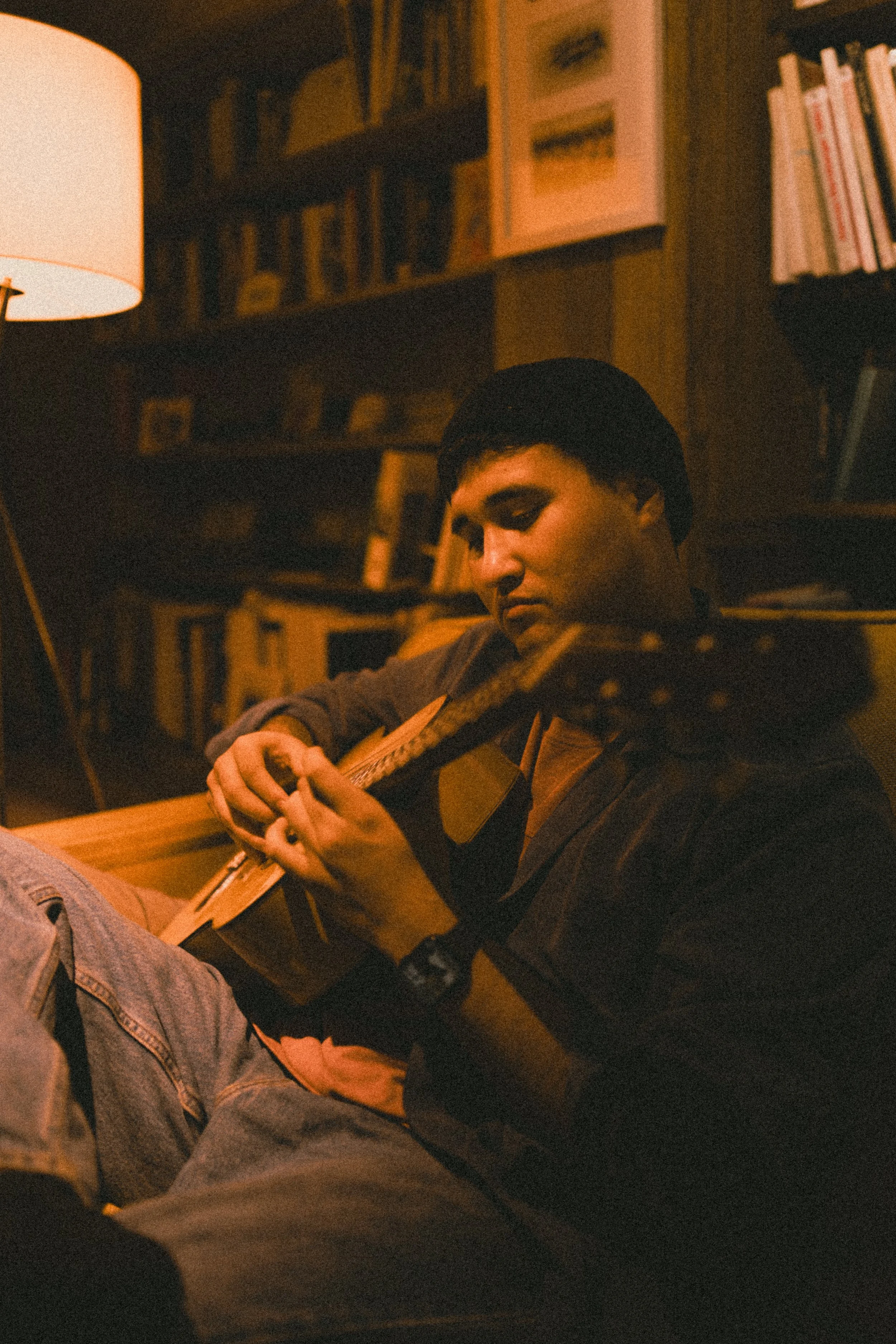 A young man playing an acoustic guitar indoors, sitting on the floor near a bookshelf and a warm-colored lamp.