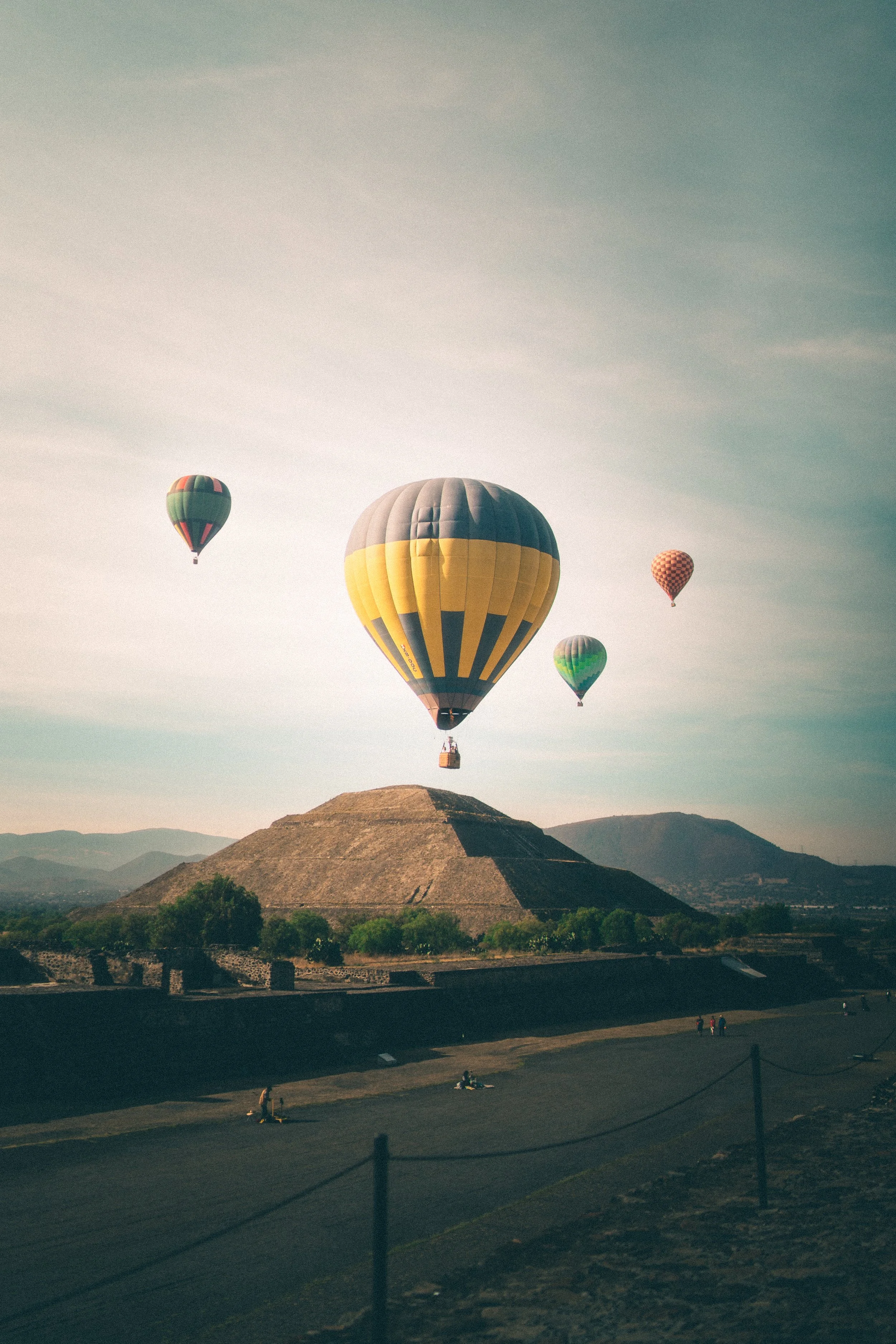 Hot air balloons floating in the sky above a historic pyramid-shaped building with trees and mountains in the background.