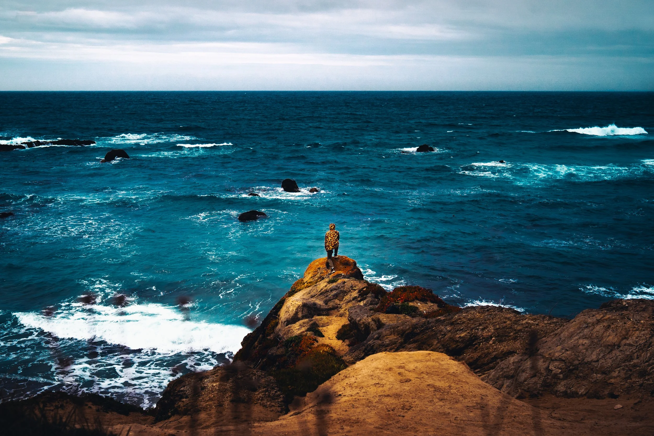 Person standing on a rocky cliff overlooking the ocean with waves crashing below and an overcast sky