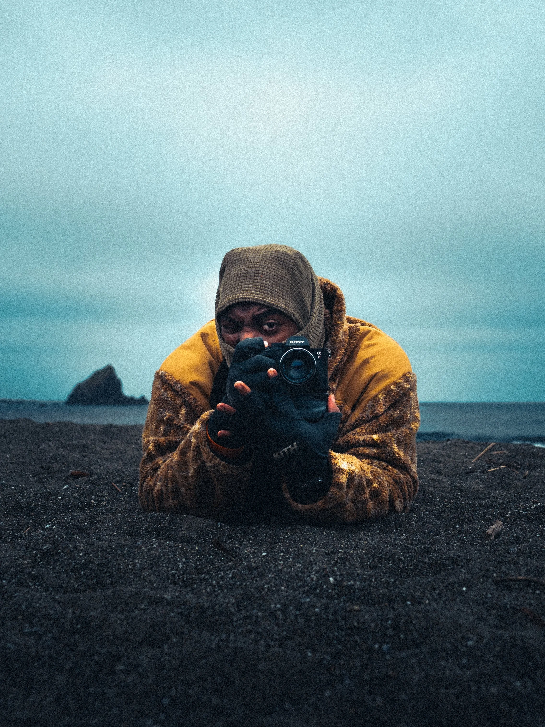 Person lying on dark sand at the beach, taking a photo with a Sony camera, wearing a brown beanie, yellow jacket, and black gloves, with cloudy sky and ocean in the background.