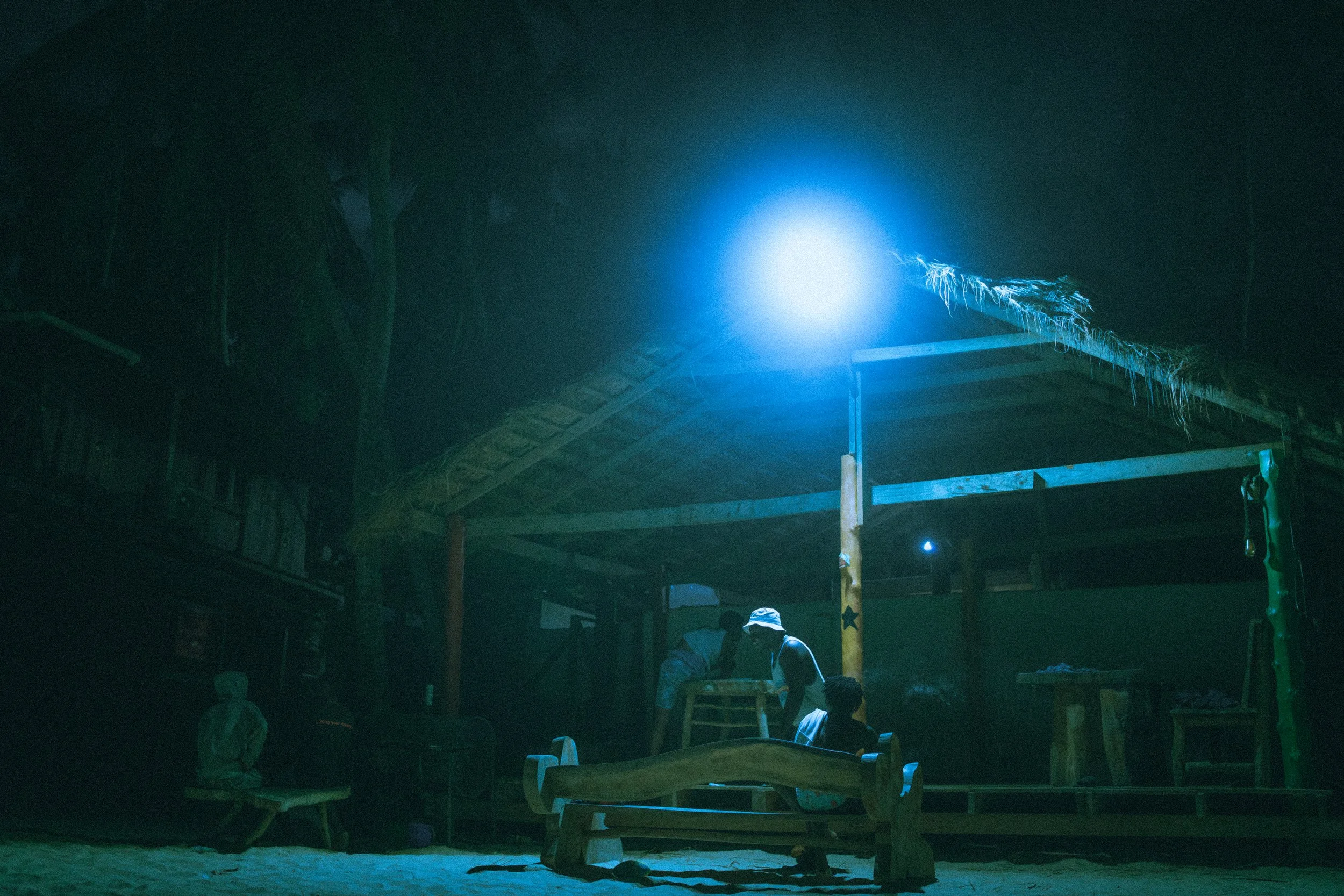 Nighttime scene of a small outdoor shelter with wooden benches and a thatched roof illuminated by a bright blue light. Two men sit under the shelter, one wearing a hat, with trees and a wooden fence in the background, creating a peaceful, rustic atmo