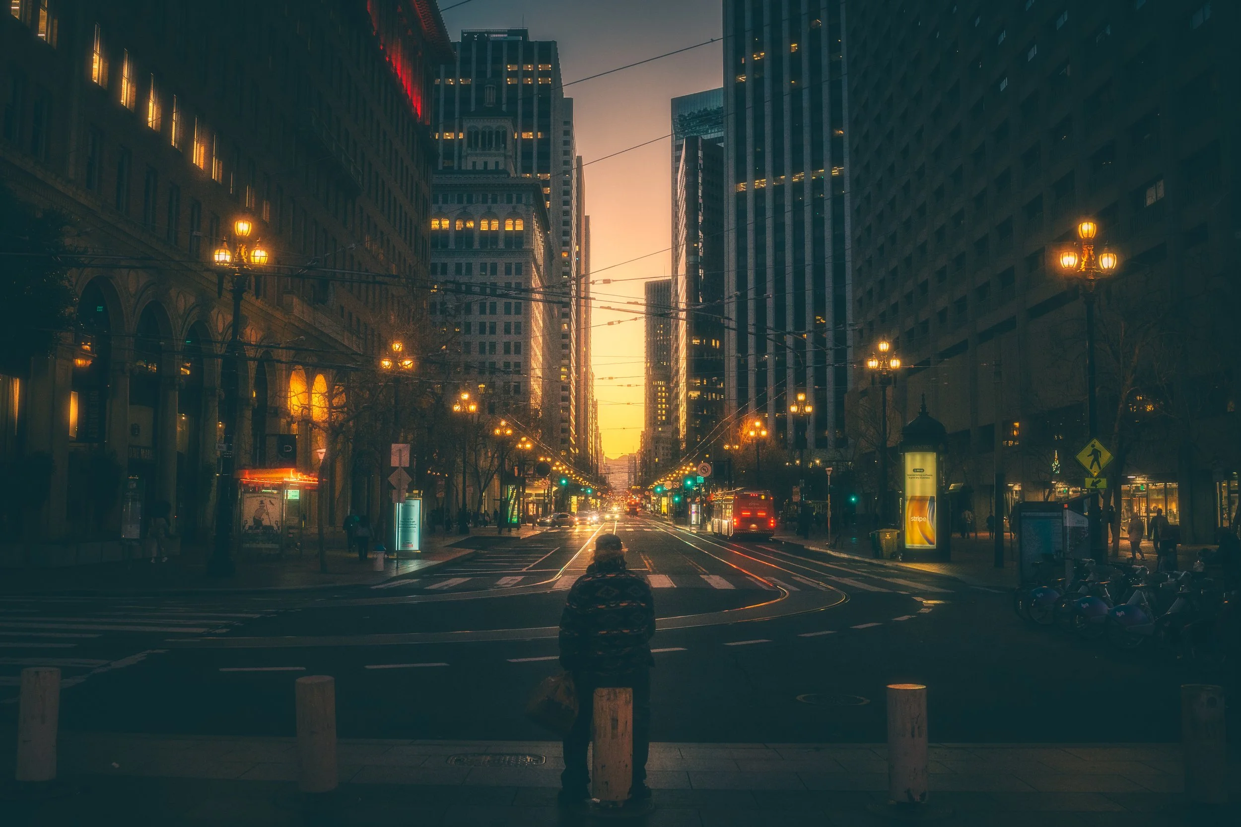 City street at sunset with tall buildings, street lamps, and tram tracks, with a solitary person standing on the curb.