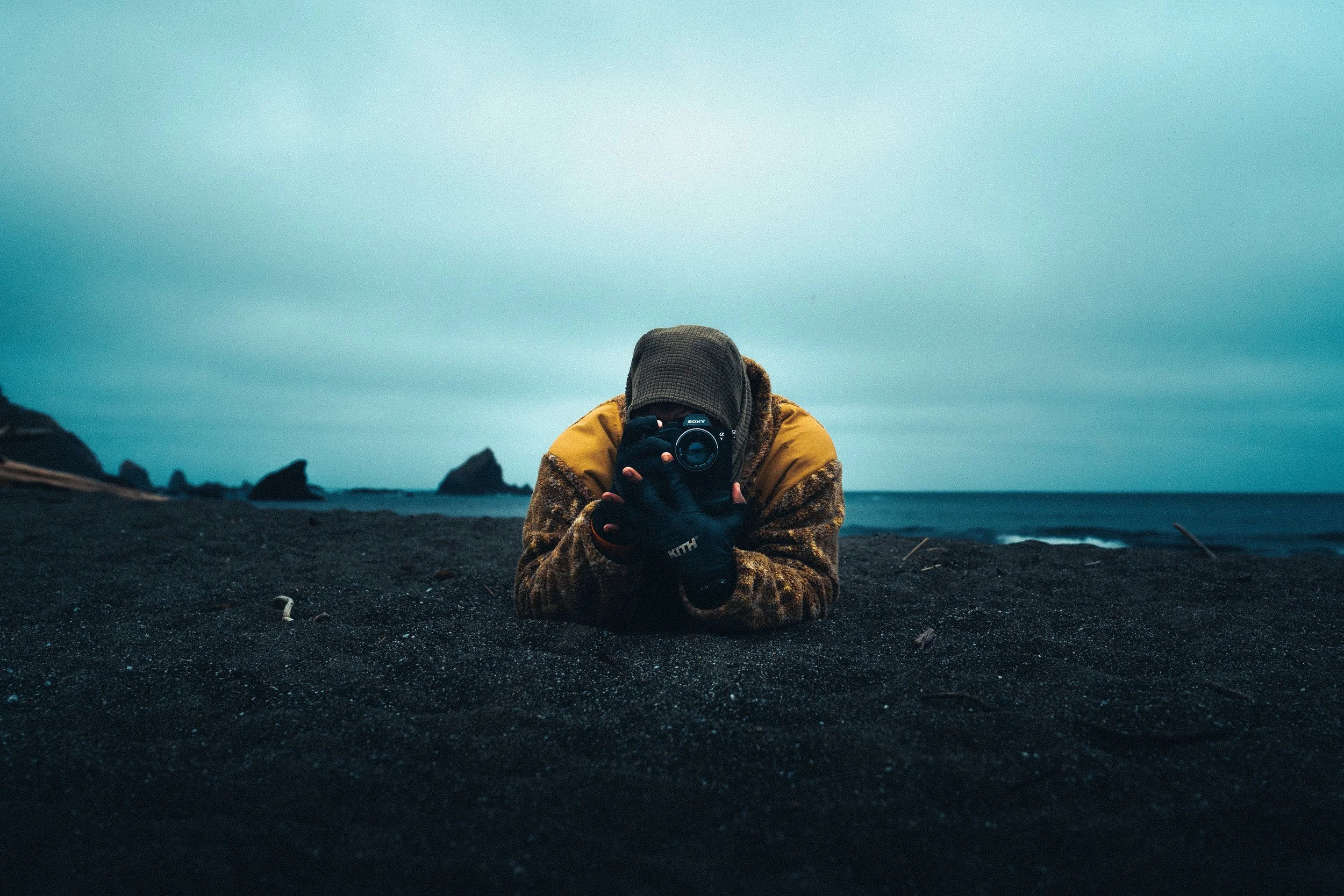 Person lying on dark sand beach taking a photo with a camera, ocean waves and rocks in the background, overcast sky.