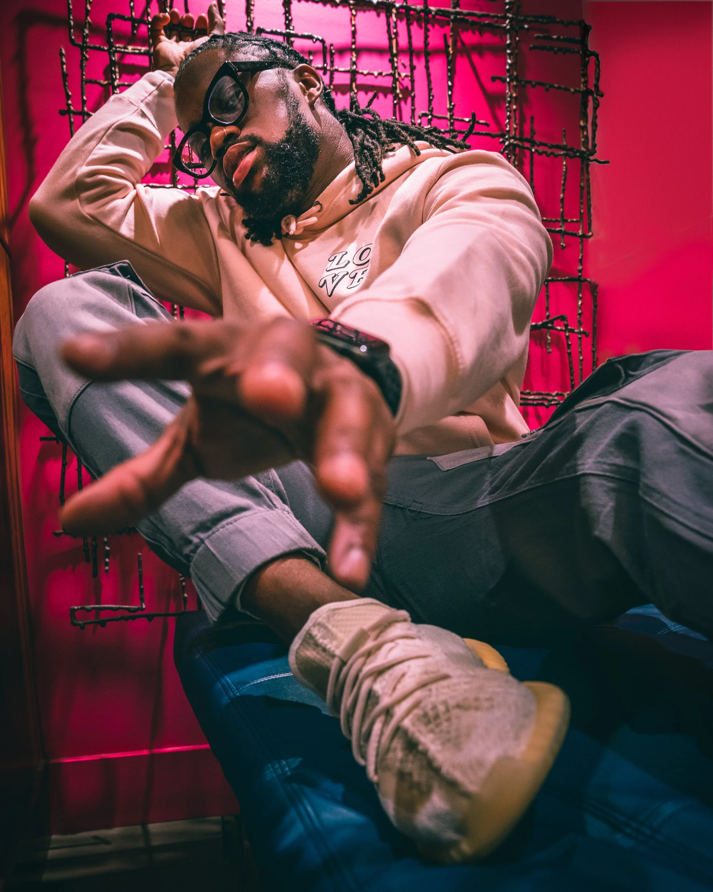 A young man with glasses and dreadlocks sitting on a bed, making a peace sign towards the camera, with a pink wall and a decorative metal grid behind him.