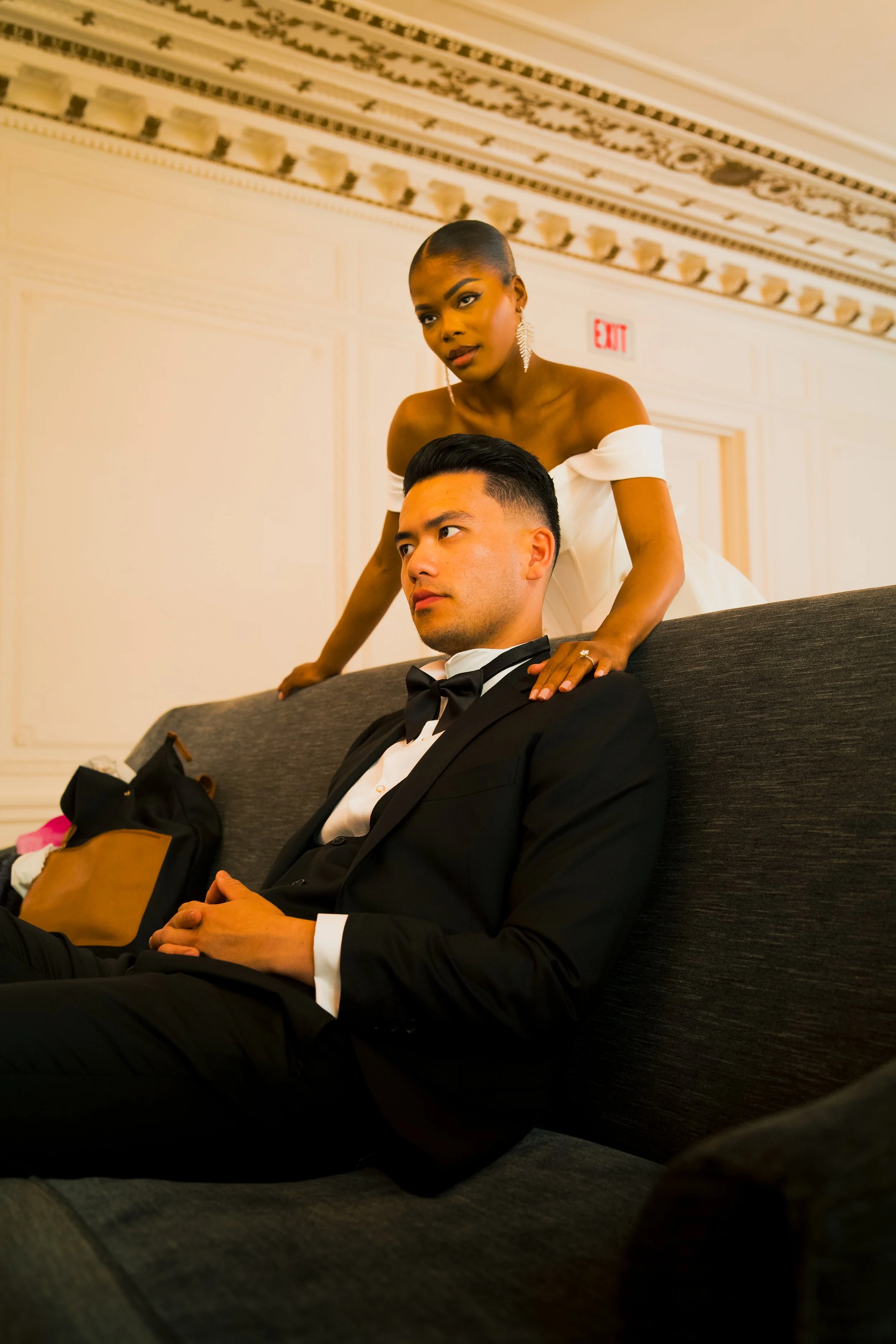 A woman in a white off-shoulder dress standing behind a man in a black tuxedo sitting on a grey sofa in an elegant room.