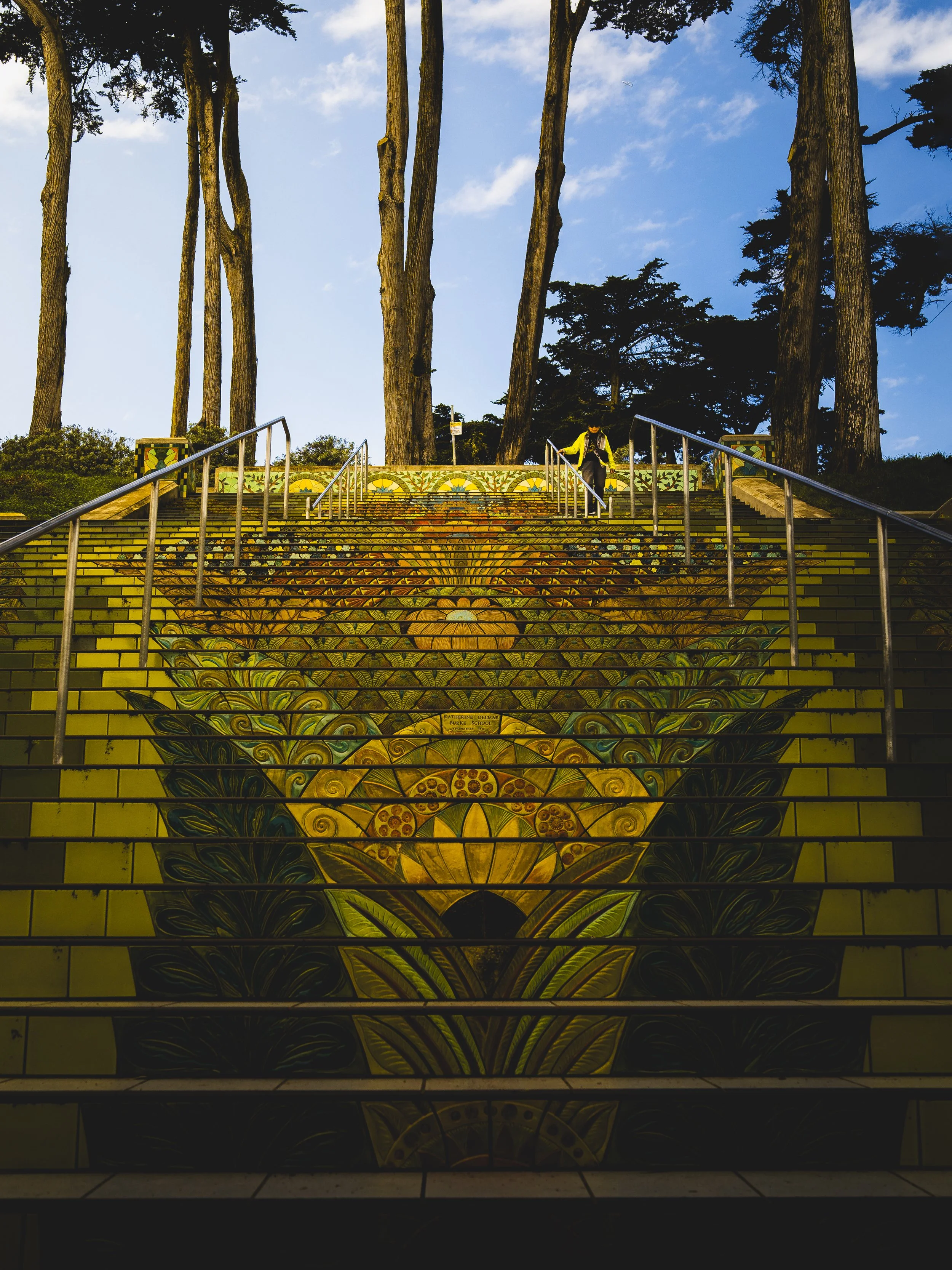 Colorful painted staircase outdoors with floral and geometric designs, set against a sky with clouds and surrounded by tall trees.