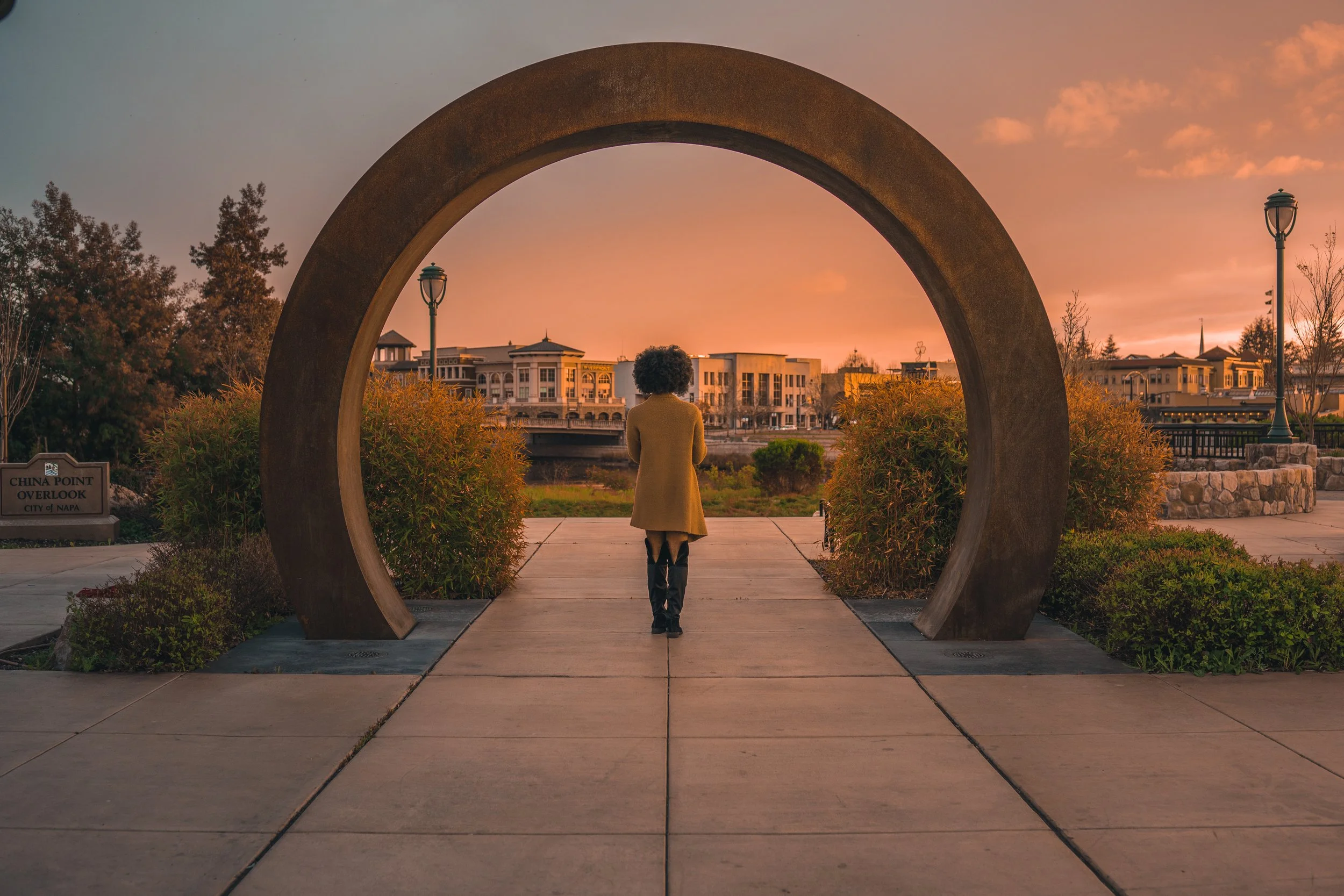 A woman walking through a circular archway in a park at sunset, with buildings and trees in the background.