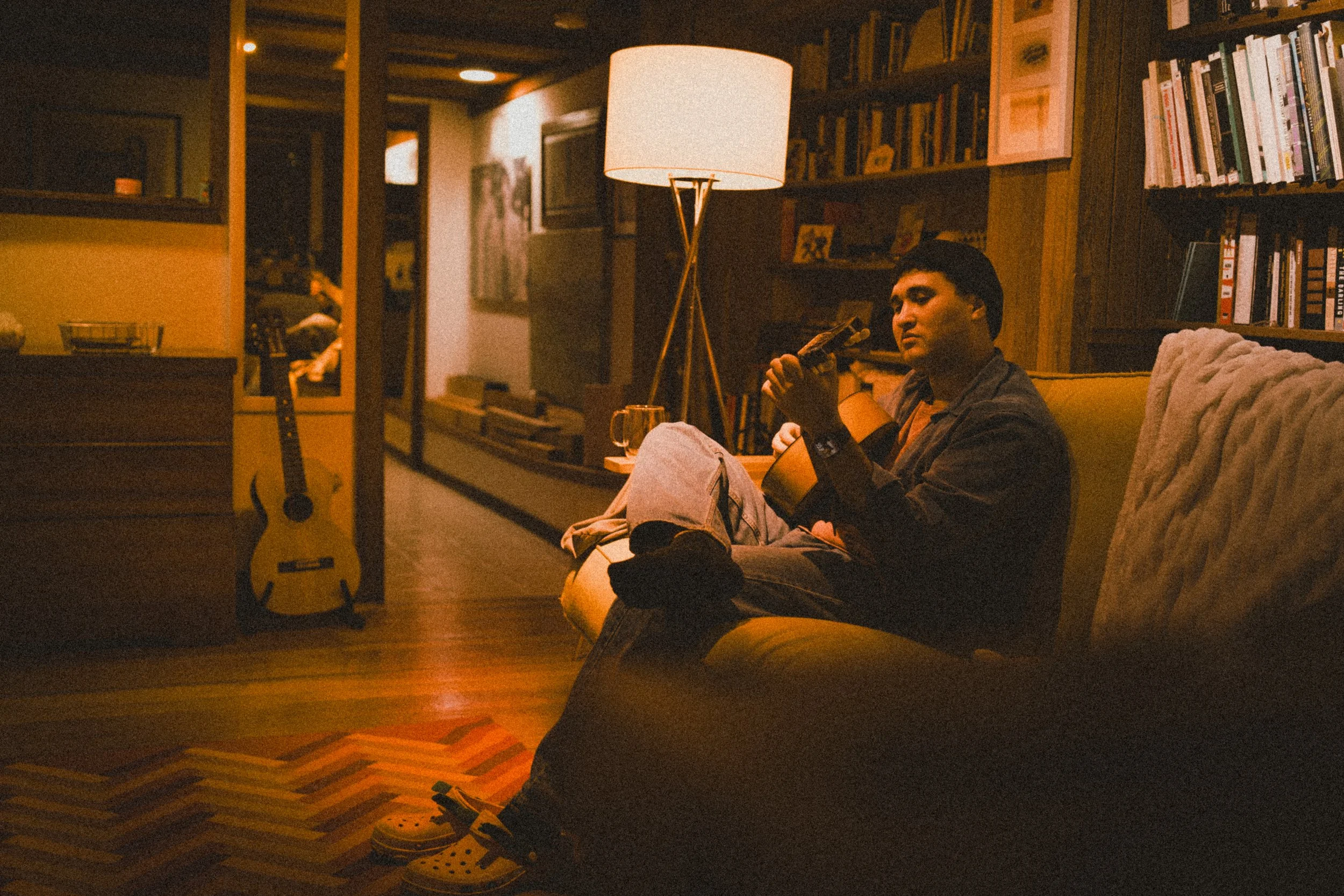 A man sitting on a yellow couch in a warmly lit living room, playing an acoustic guitar, with a drink on the side table. The room has bookshelves and a large lamp, with a guitar resting against a dresser.