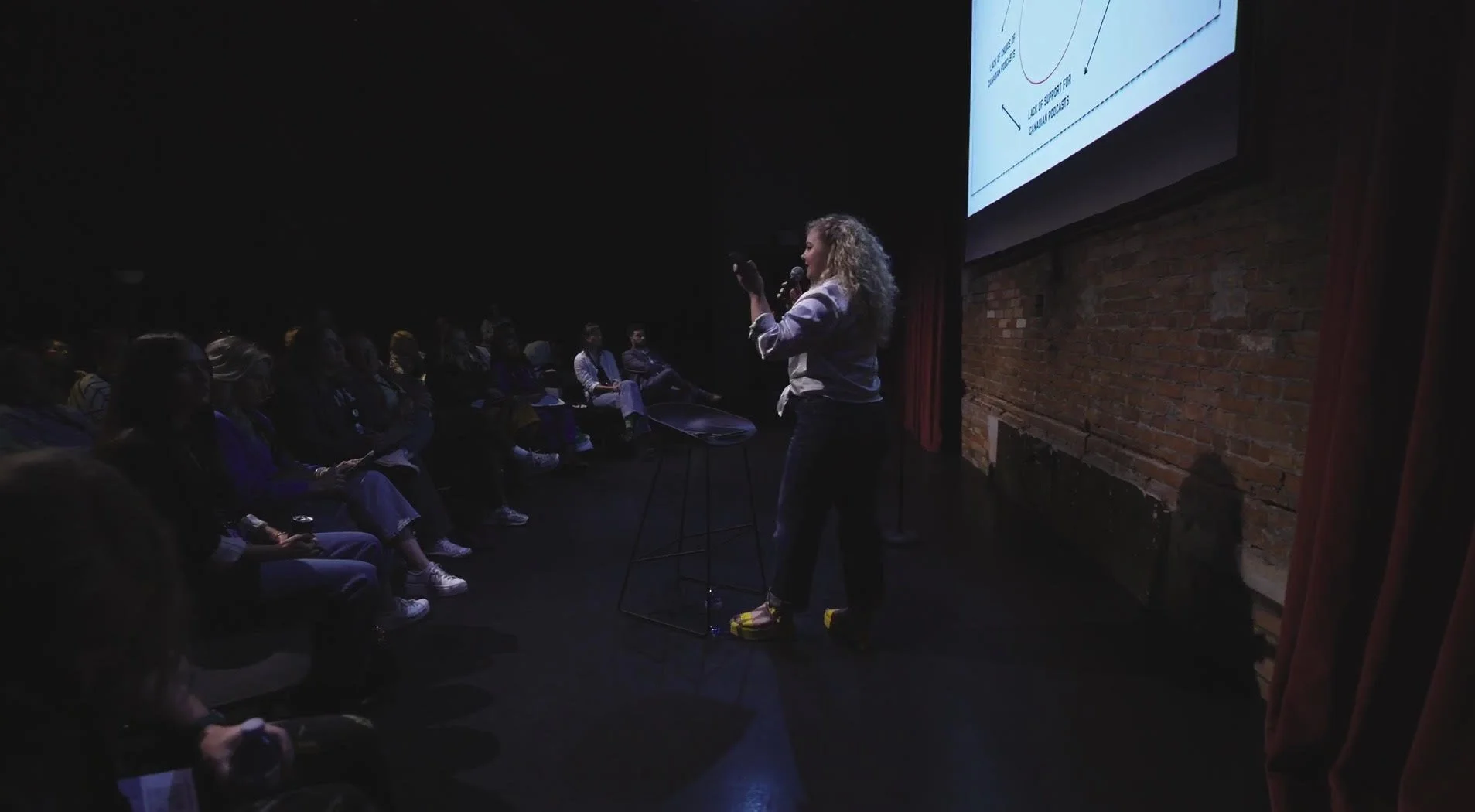 Kattie Laur giving a presentation to an audience in a dark room, with a large screen behind her projecting a graph.