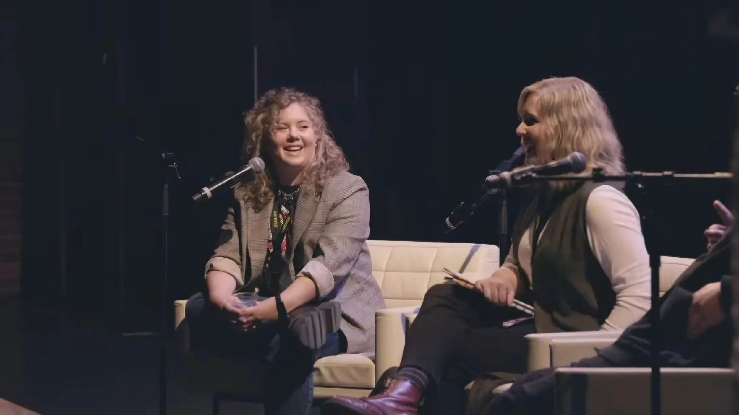 Two women sitting on a cream armchair, smiling and talking into microphones during a panel discussion or interview.