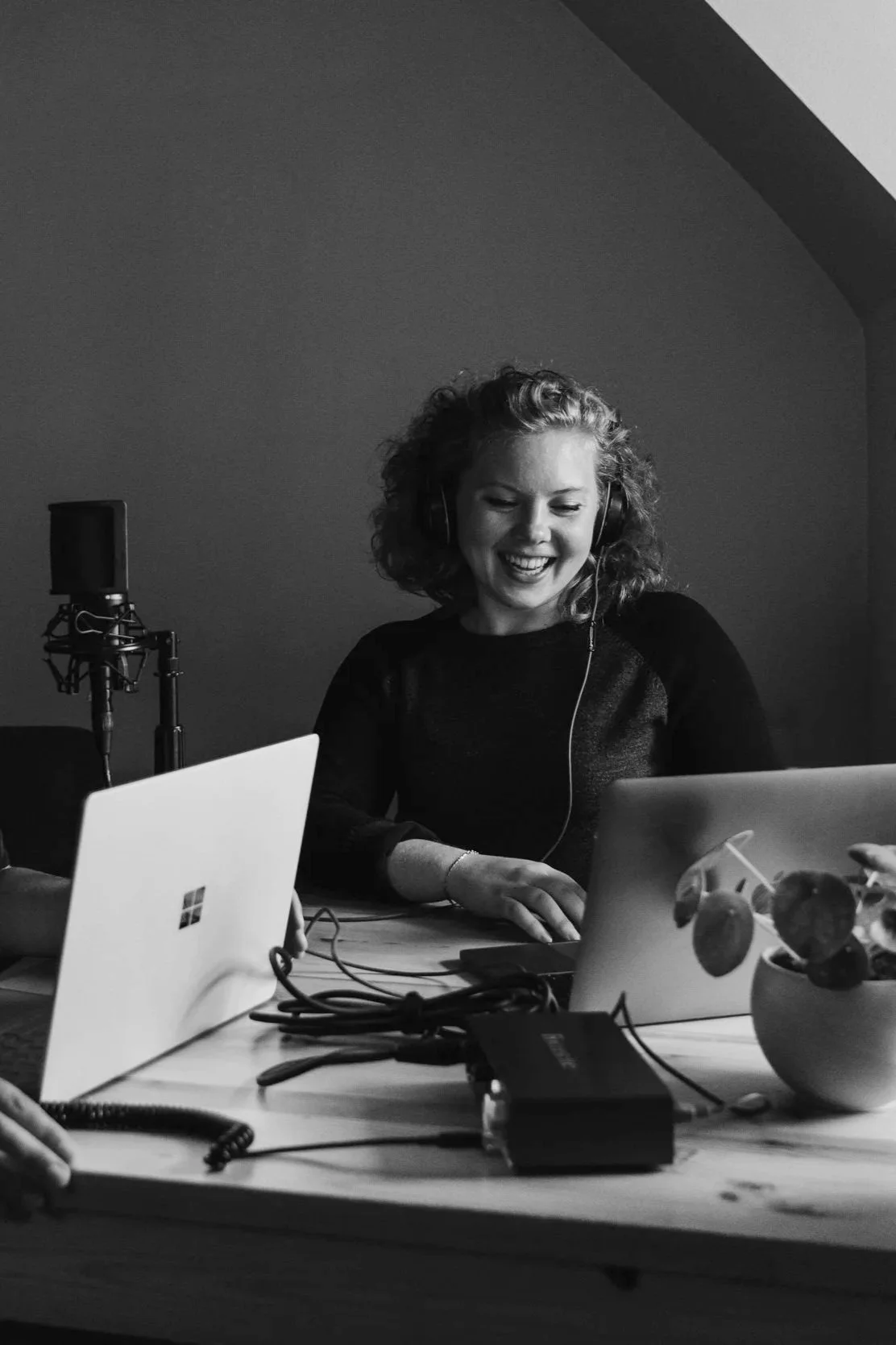 A woman wearing headphones, smiling while working on a laptop in a room with a microphone and audio equipment on a wooden table.