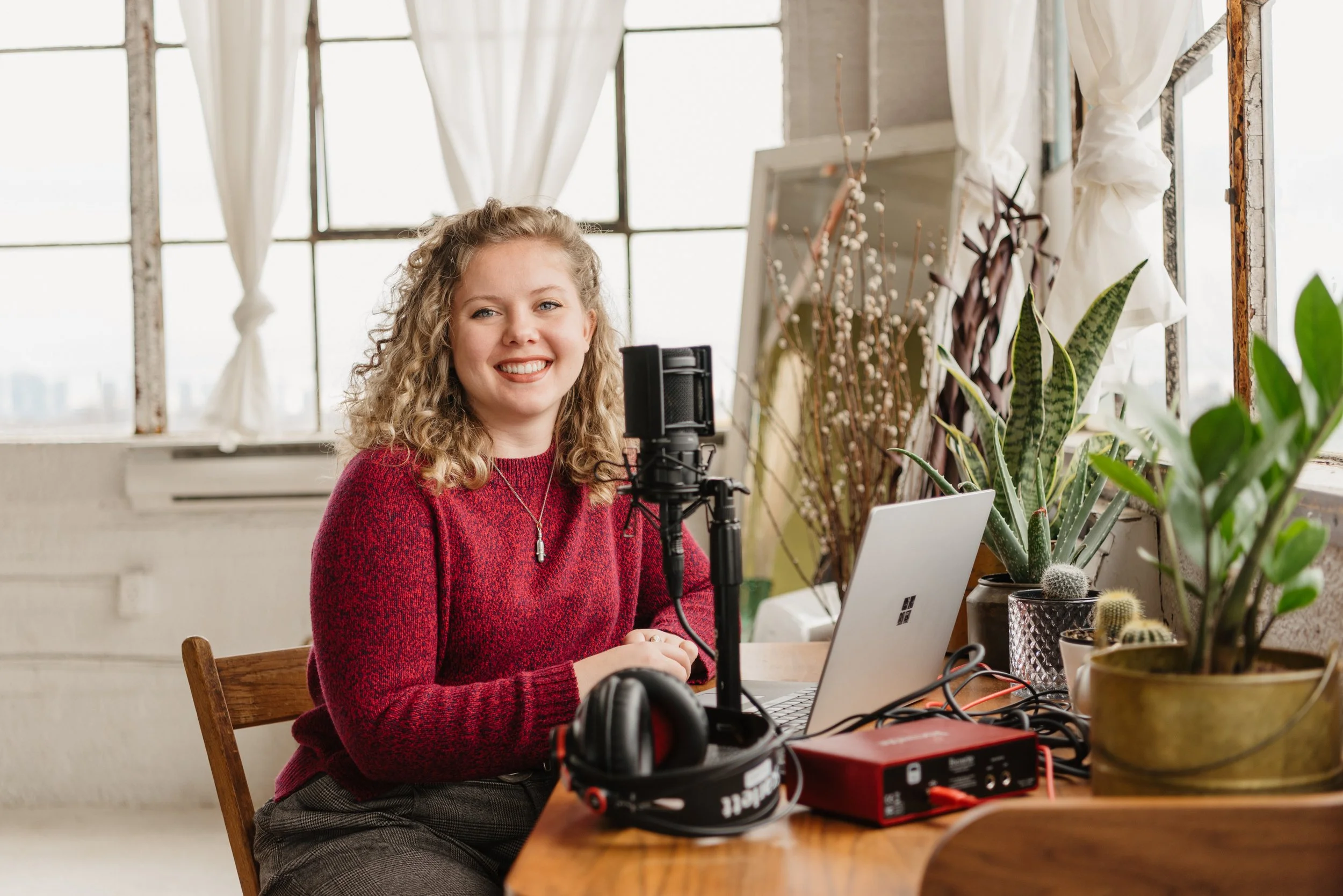 A woman with curly blonde hair wearing a red sweater, sitting at a wooden table with a laptop, microphone, headphones, and recording equipment, smiling in a bright room with large windows and plants.