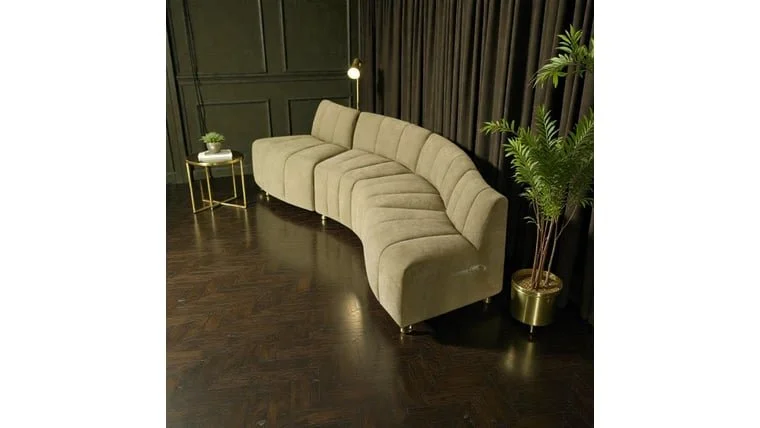 A beige sectional sofa in a room with dark wood floors, accompanied by a small side table with a potted plant, and a tall indoor plant in a gold pot next to dark curtains.
