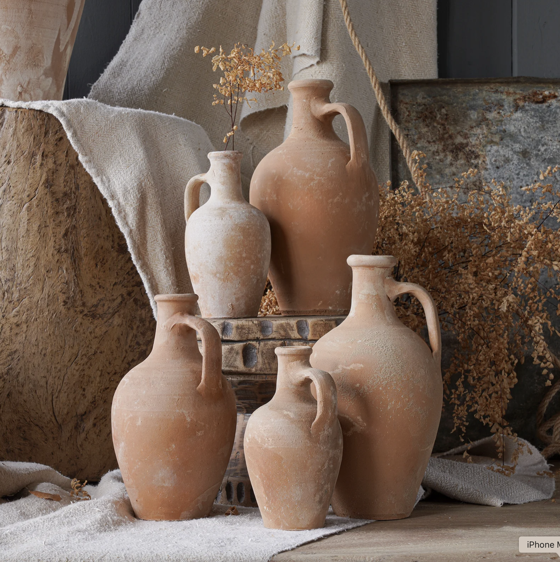 Several ancient-style clay vases in various sizes and shapes arranged on a table with a rustic backdrop, brown dried foliage, and textured fabric.