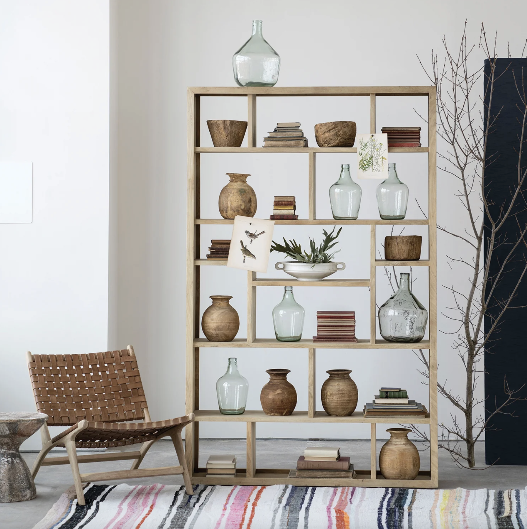 Wooden bookshelf with glass vases, pottery, and books, a brown woven chair, a striped rug, and a leafless tree branch in the background.