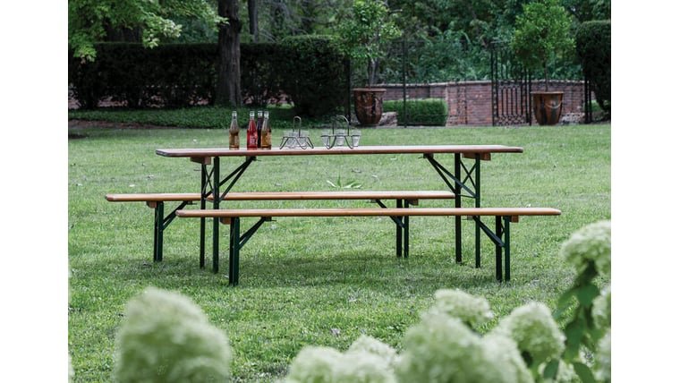 Outdoor picnic table with condiments and decor on a grassy lawn, surrounded by trees and garden elements.
