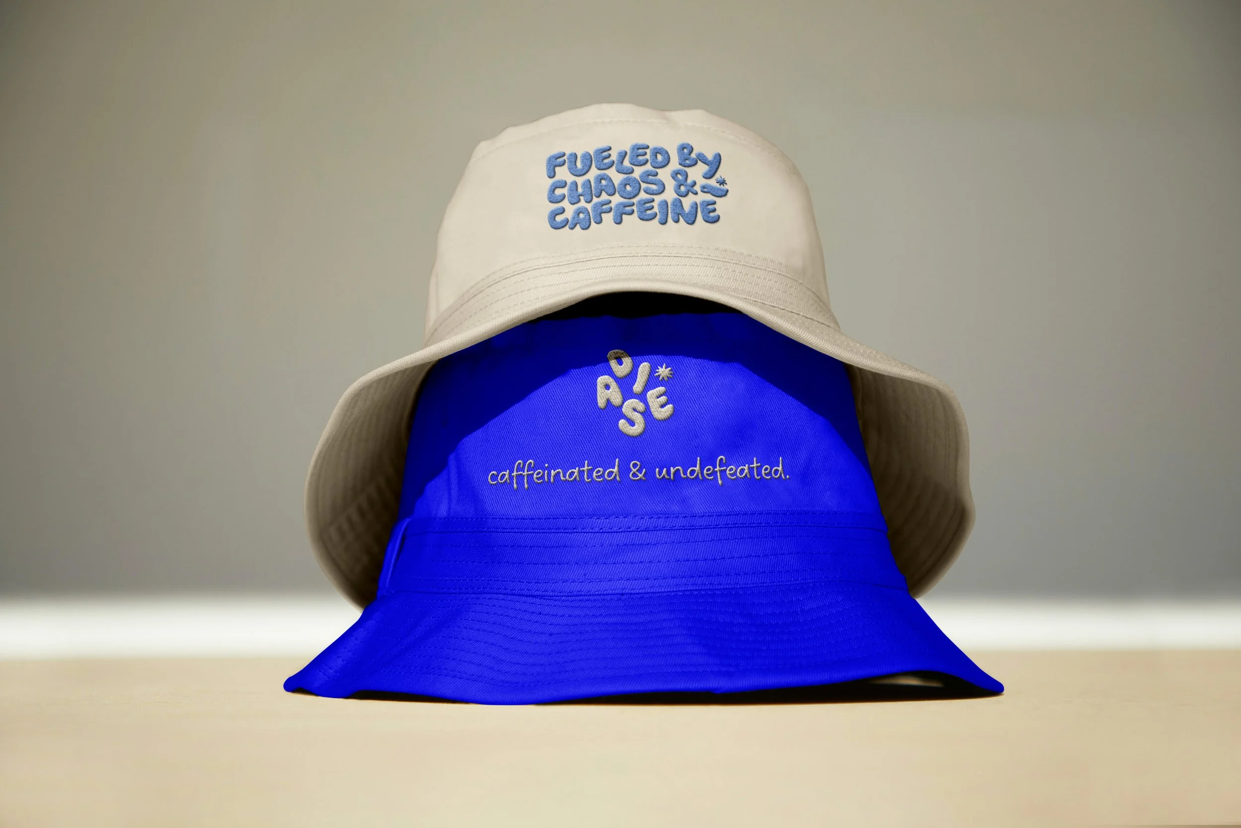 Two bucket hats, one beige and one blue, stacked with the beige on top. The beige hat has embroidered text in blue that says, 'Fueled by chaos & caffeine.' The blue hat has embroidered white text that says, 'R.O.S.E caffeineed & undefeated.'
