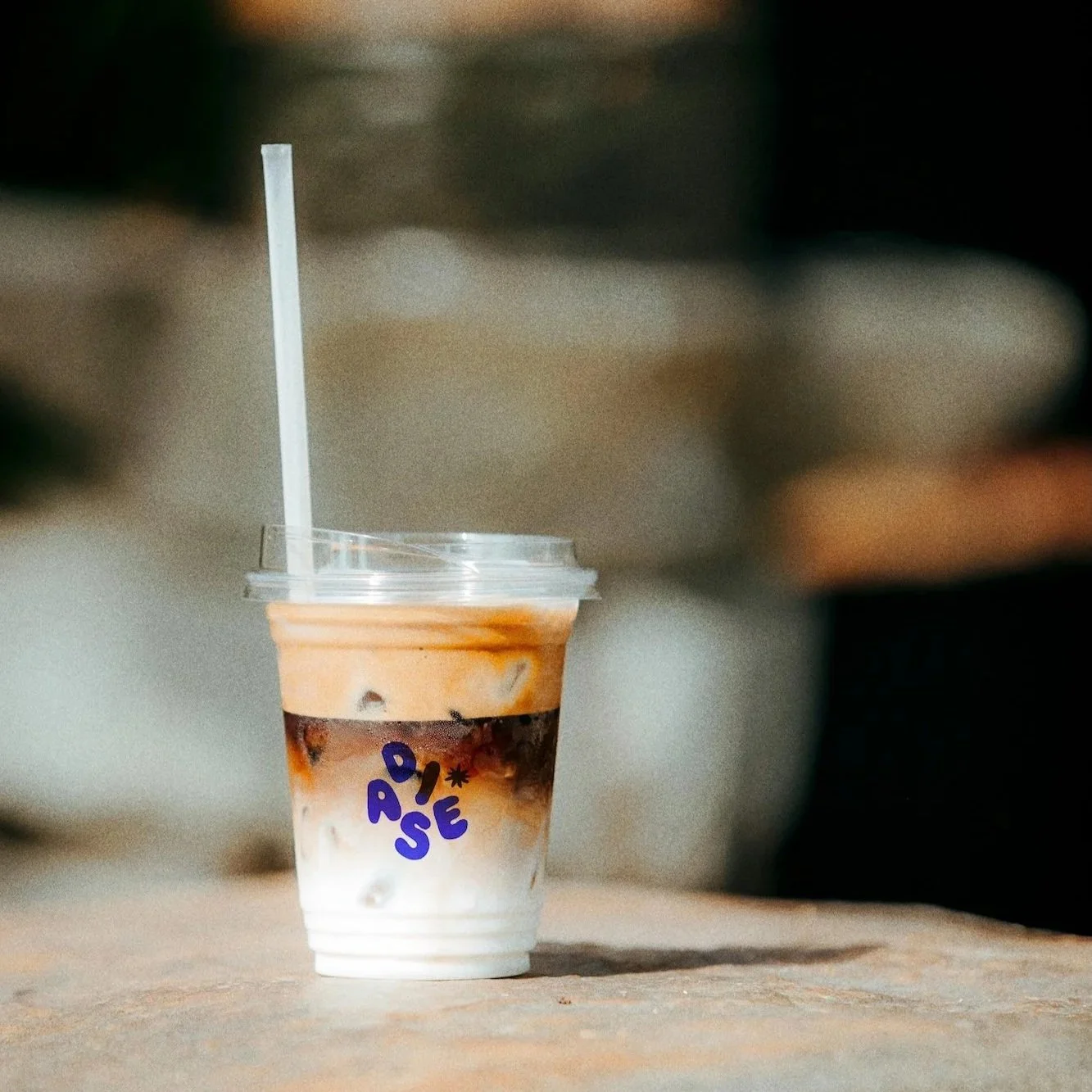 A plastic cup with a clear lid and straw, containing iced coffee with layers of milk and coffee, sitting on a concrete surface.