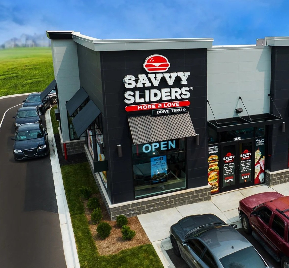 Fast food restaurant building with a sign that reads "Savvy Sliders" and "More 2 Love," with cars parked in front and background of a grassy landscape under a blue sky.