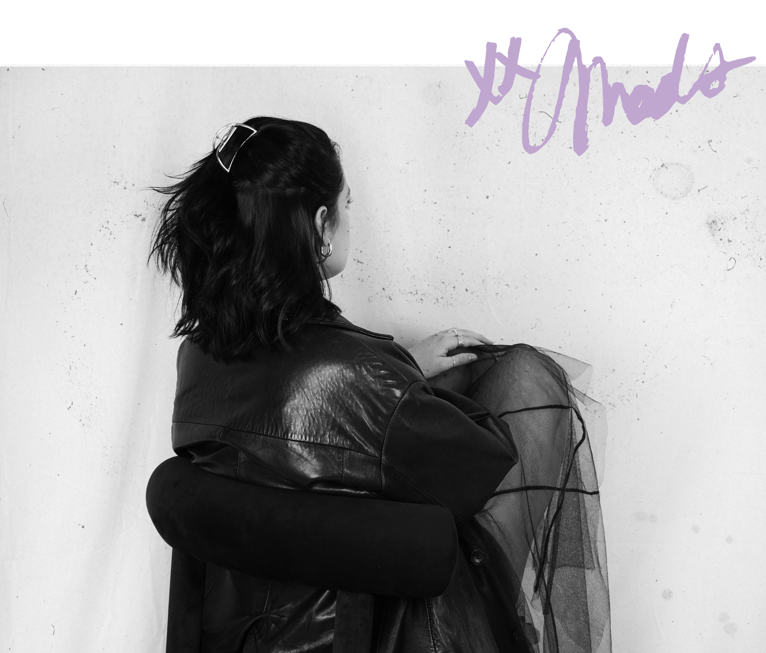 Black and white photo of a woman sitting on a chair, facing a wall, wearing a leather jacket and hoop earrings, with loose shoulder-length hair, and a hair clip.