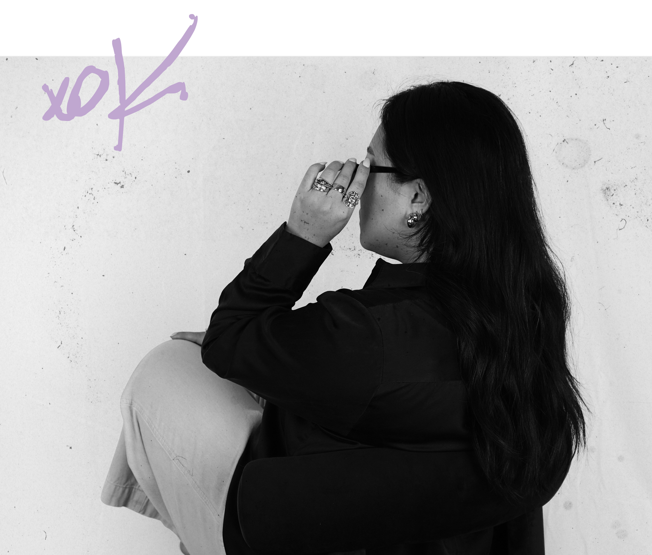 A woman with long dark hair and glasses, wearing jewelry, sits with her knees up and hair covering her face, against a white textured wall with handwritten purple text saying 'xoxo' in the upper left corner.