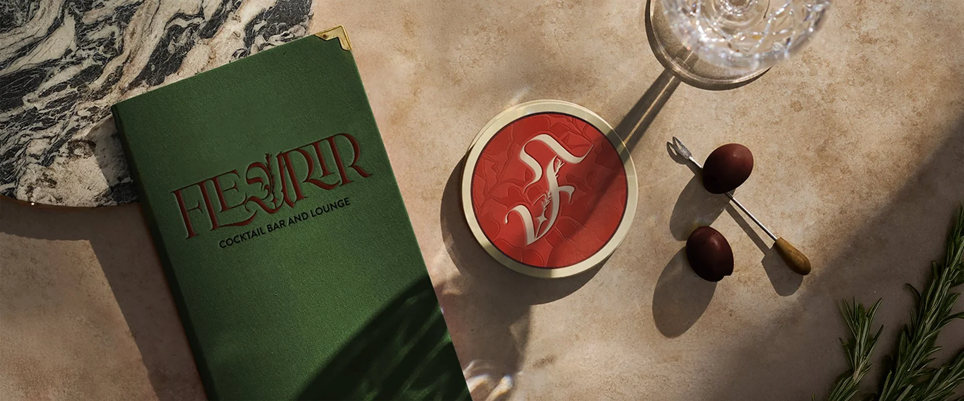 Objects on a bar counter including a green menu for 'Fleurir Cocktail Bar and Lounge', a red circular container with the bar logo, a martini glass, a small fork and olives, and a sprig of rosemary.