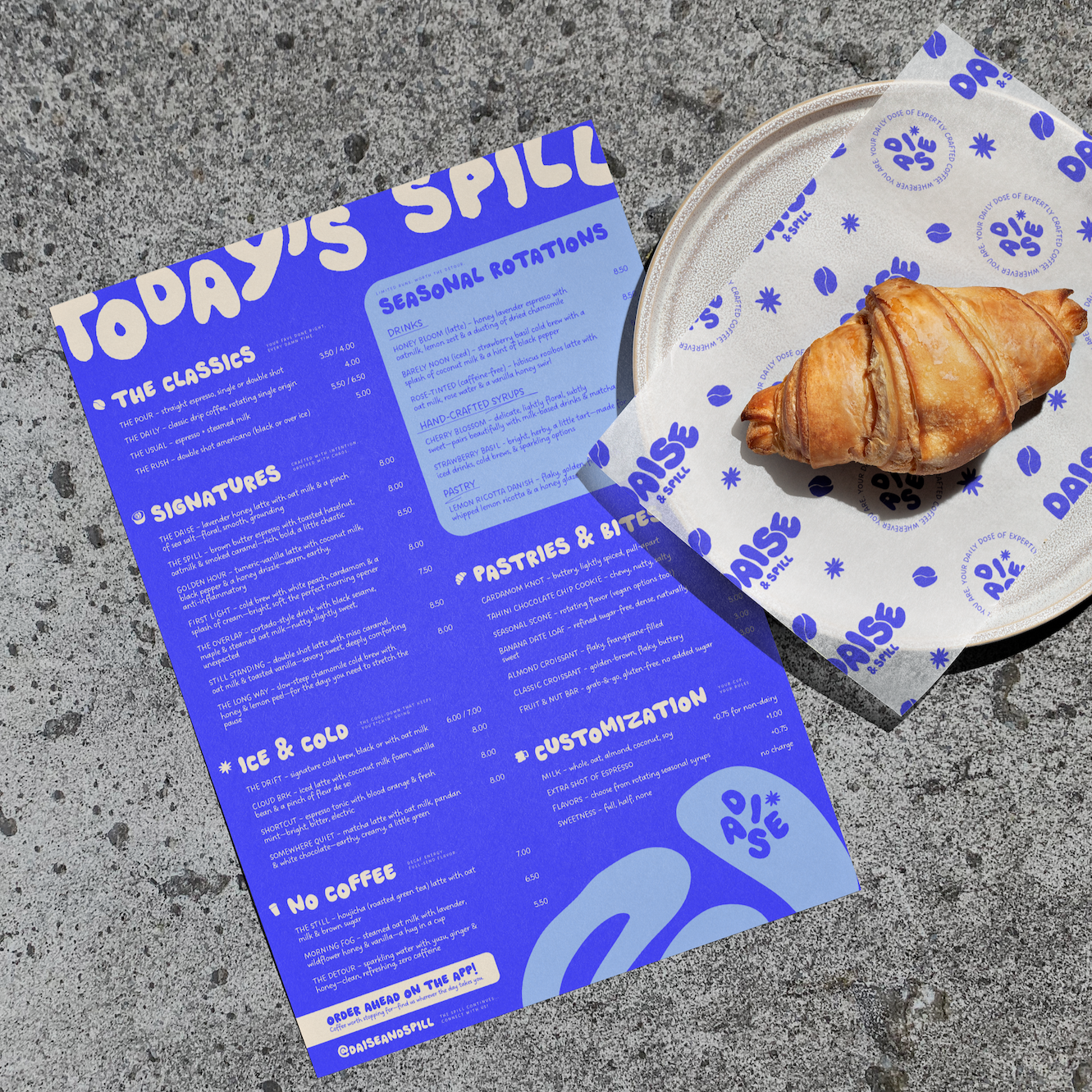 Blue menu paper with white text listing drinks, pastries, and customizations, placed next to a croissant on a tray lined with branded paper at a cafe.