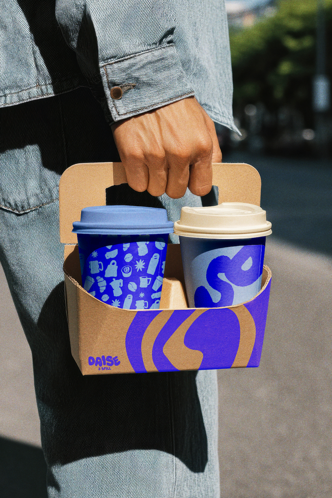 A person's hand holding a cardboard tray with two coffee cups, one blue with a purple lid and graphic design, and the other white with a purple design, outside on a sunny day.