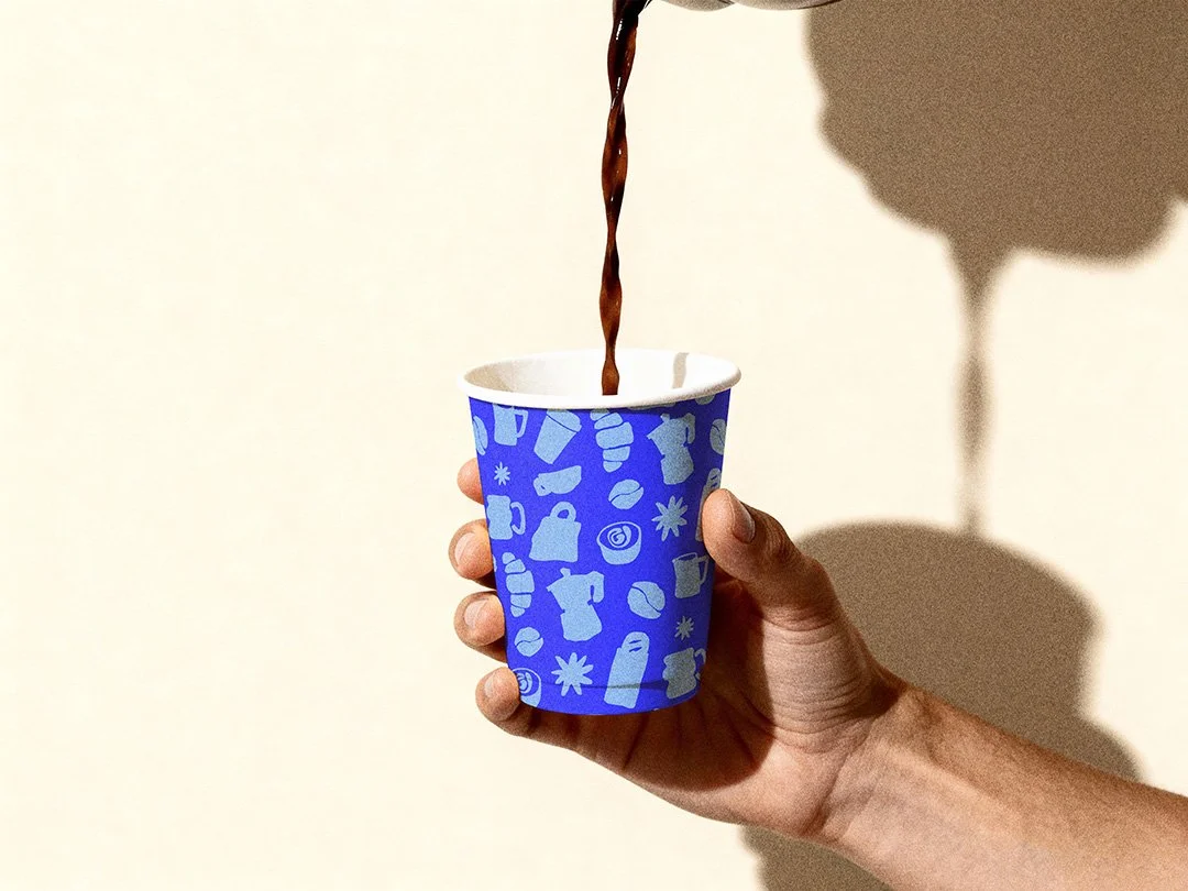 A hand holding a blue paper cup with white icons, pouring dark liquid into the cup, casting a shadow on the wall.