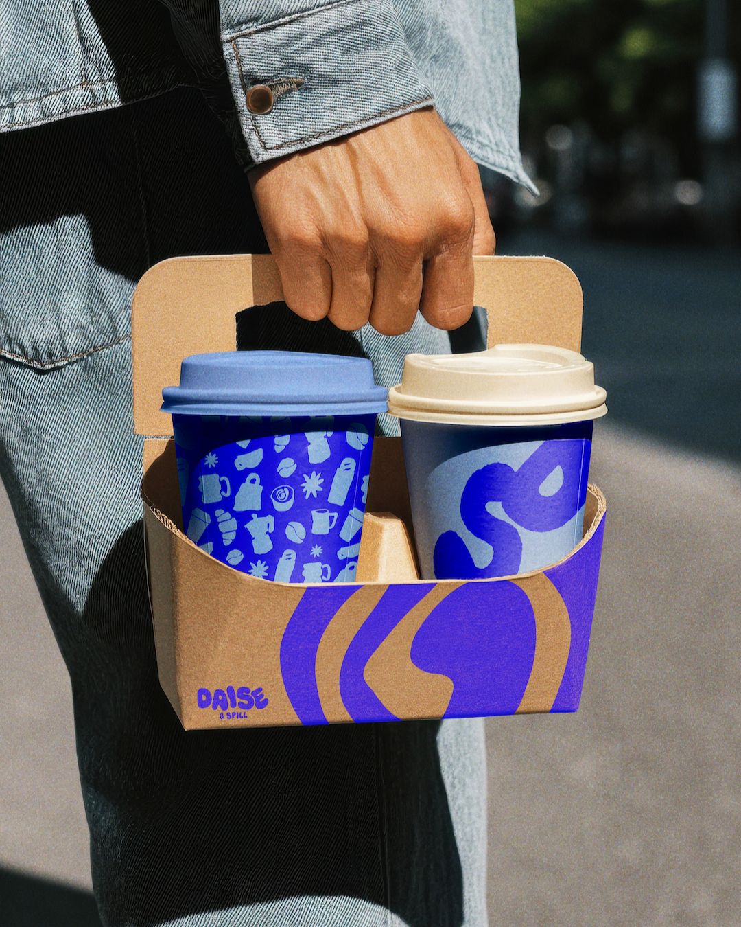 A person holding a cardboard carrier with two disposable coffee cups, one blue with a lid, and the other beige and purple with a lid, outdoors.