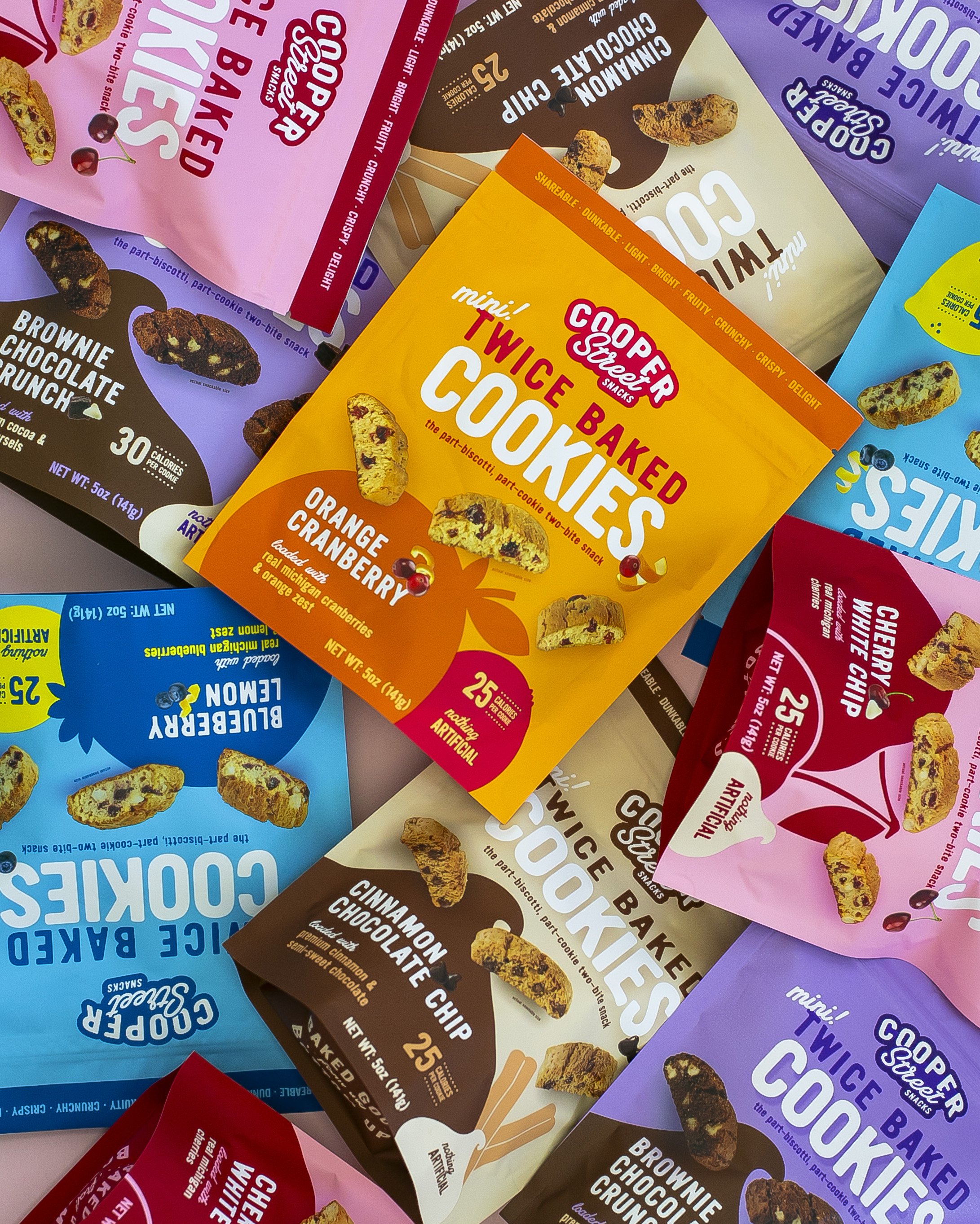 Several bags of Coop Street Snacks mini baked twice cookies, including orange cranberry, cinnamon chocolate chip, cherry white chip, and double layered cookies, arranged in a colorful display.