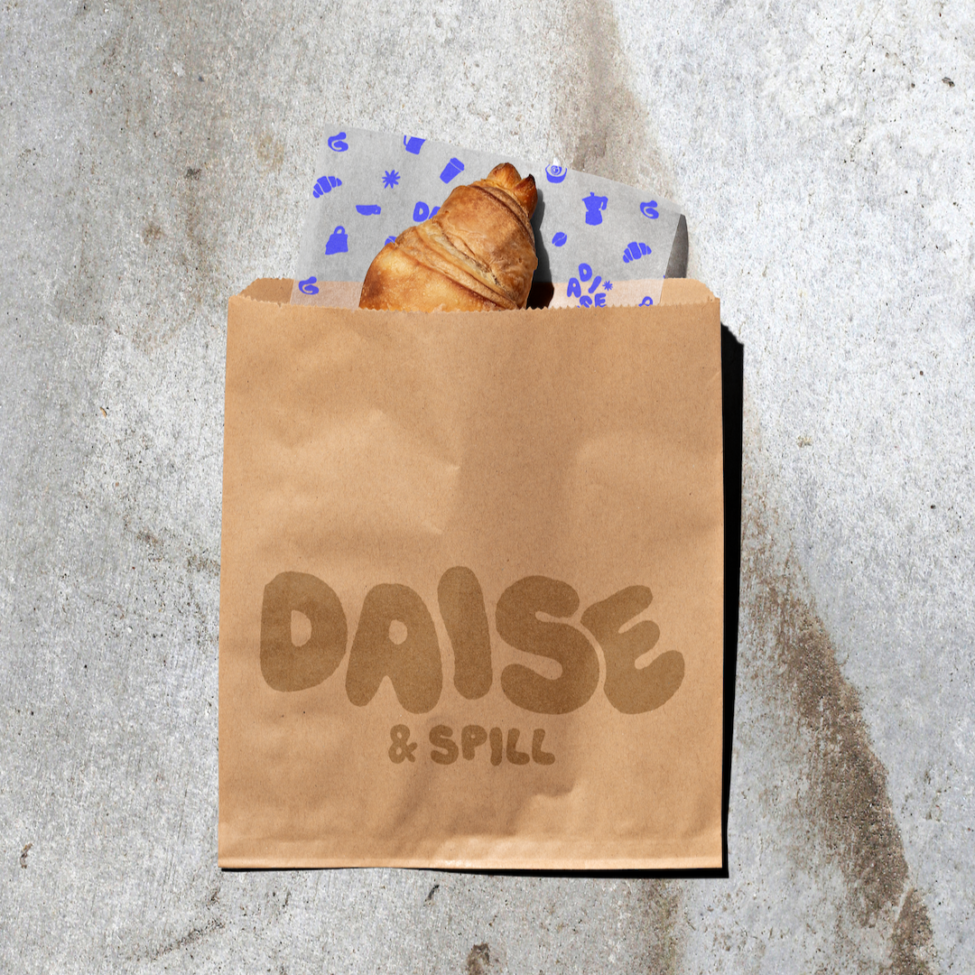 A brown paper bag labeled 'DAISE & SPILL' with a croissant inside, sitting on a concrete surface.