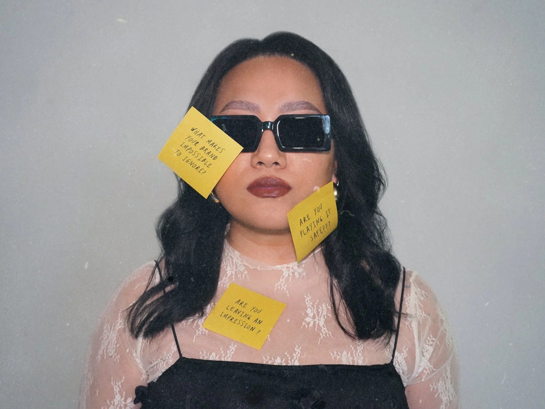 Woman with long black hair wearing black sunglasses and a lace top, with three yellow sticky notes on her face and chest, against a plain background.