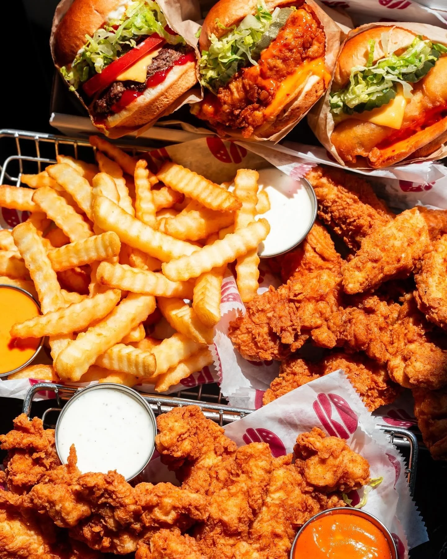 A basket of fried chicken, crinkle-cut French fries with dipping sauces, and three cheeseburgers with lettuce, tomato, cheese, and various toppings.